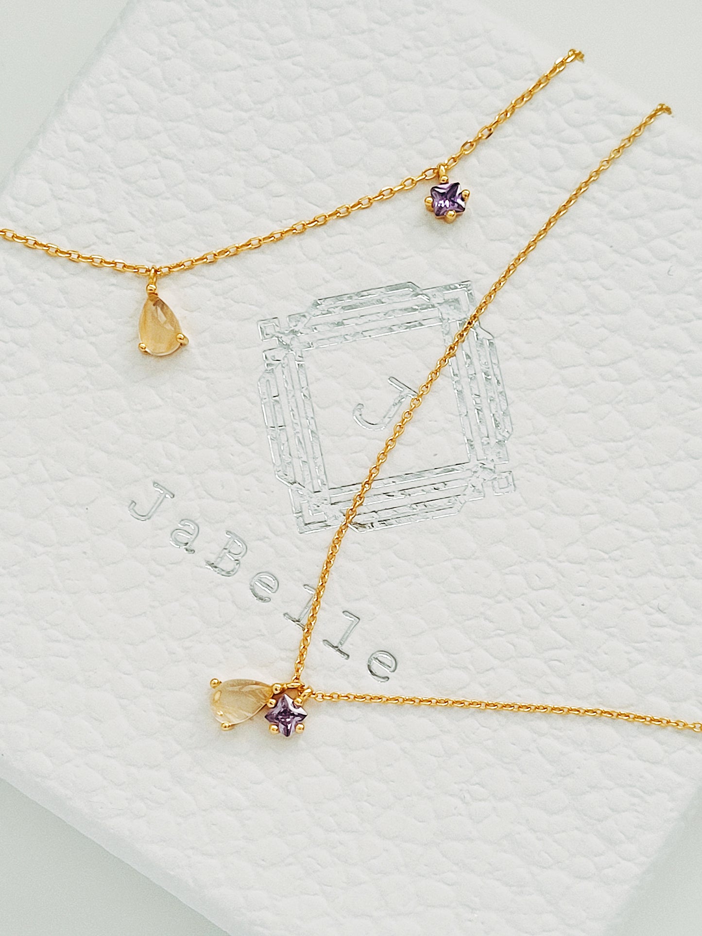 A delicate gold bracelet is displayed on a white background, featuring a teardrop-shaped rutilated quartz charm alongside a small purple amethyst star-shaped charm. The fine gold chain adds to the minimalistic elegance of the design.