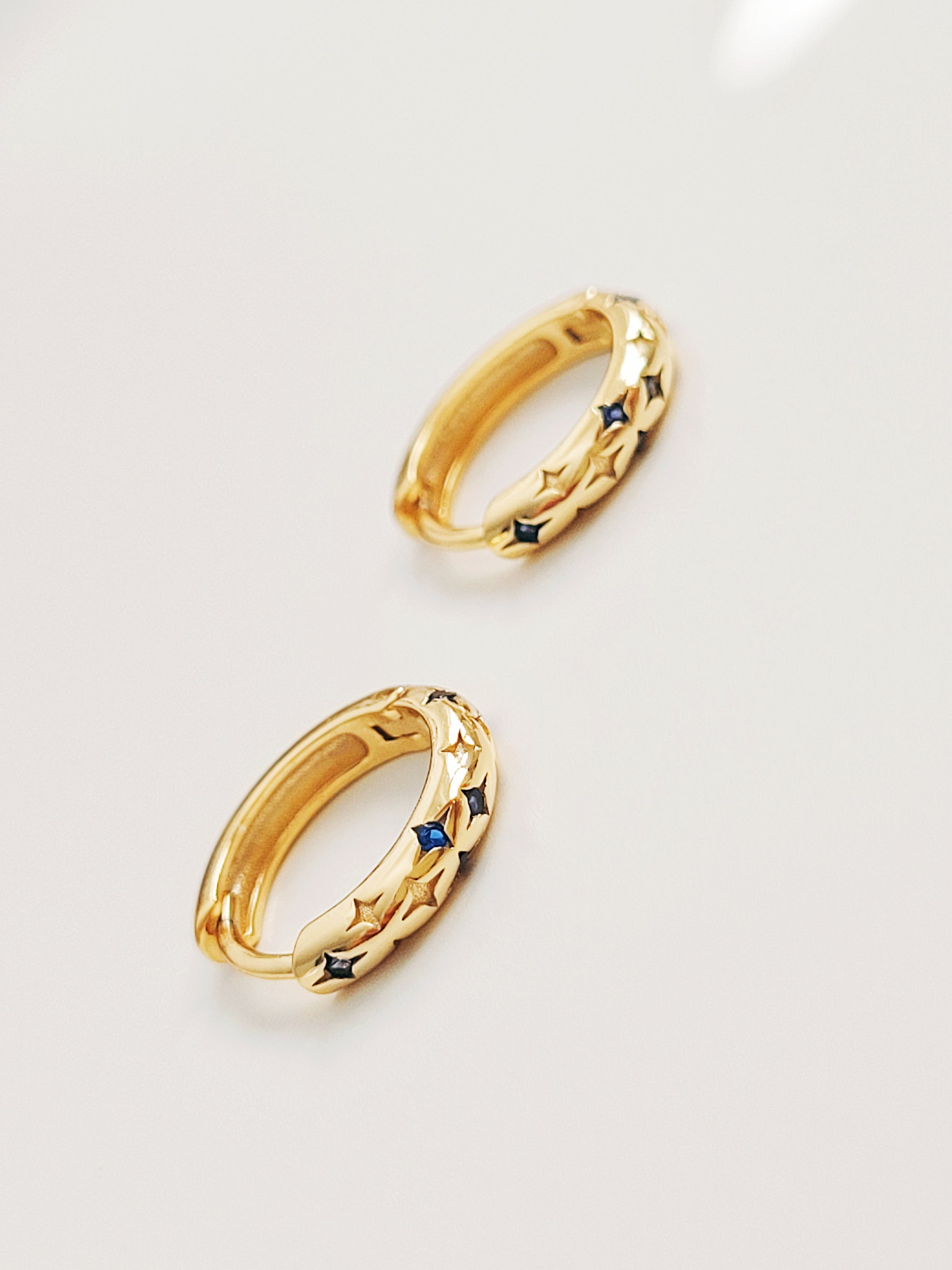 A pair of delicate huggie earrings with starry details with blue gemstone displayed on a white surface