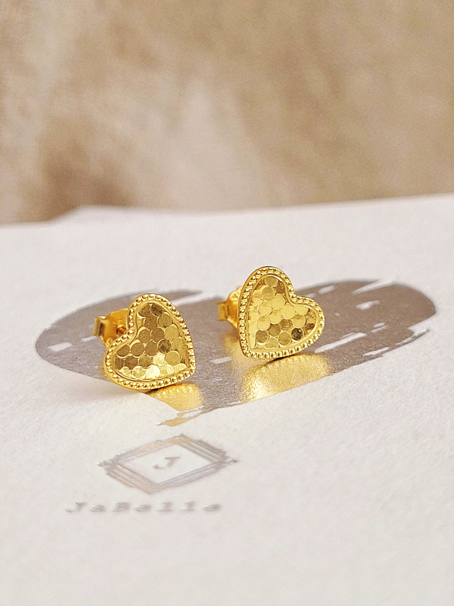 Gold heart stud earrings with scalloped texture on a white surface with a beige background