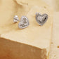 Silver heart stud earrings with scalloped texture on a white surface with a beige background