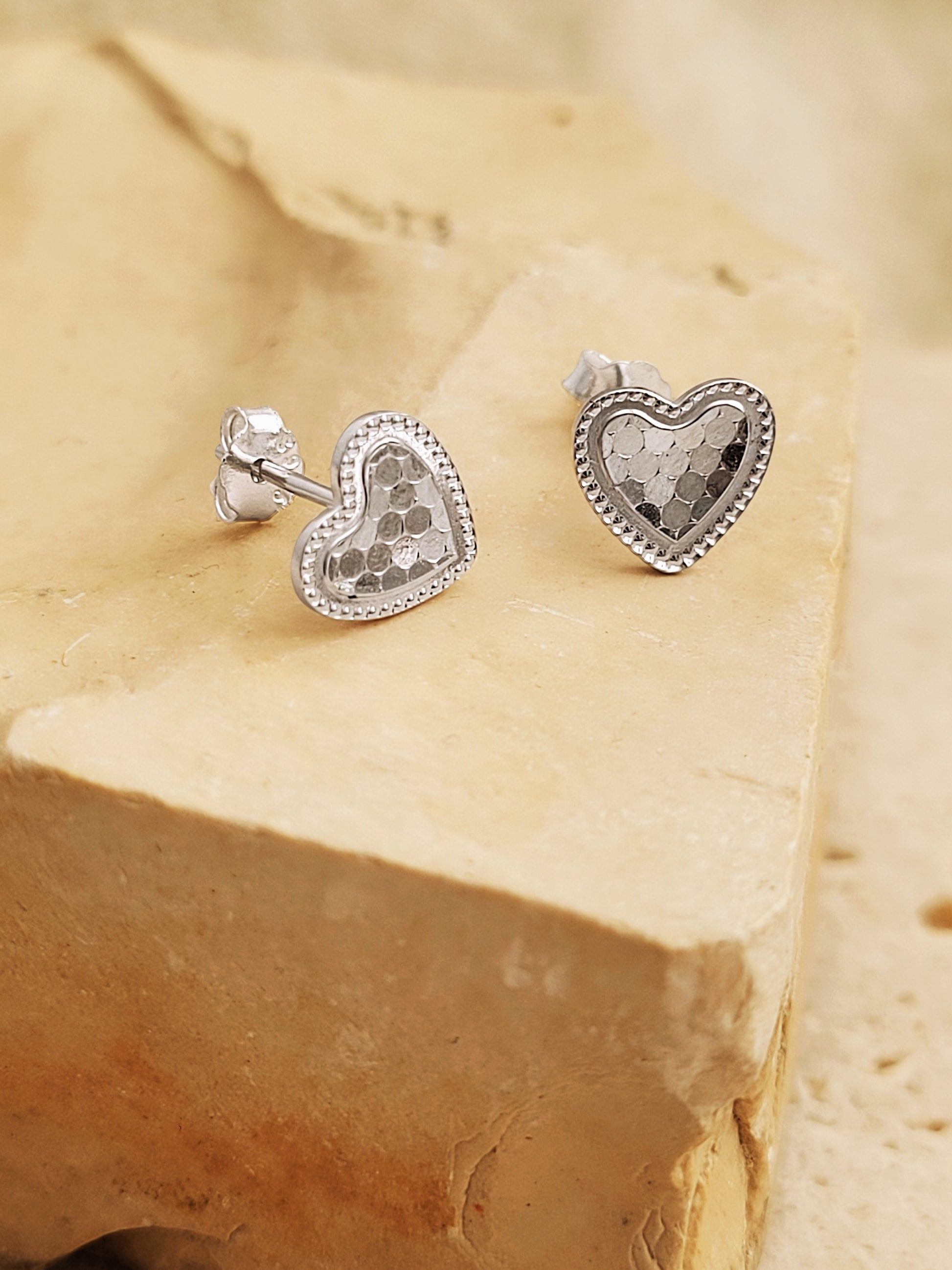 Silver heart stud earrings with scalloped texture on a white surface with a beige background