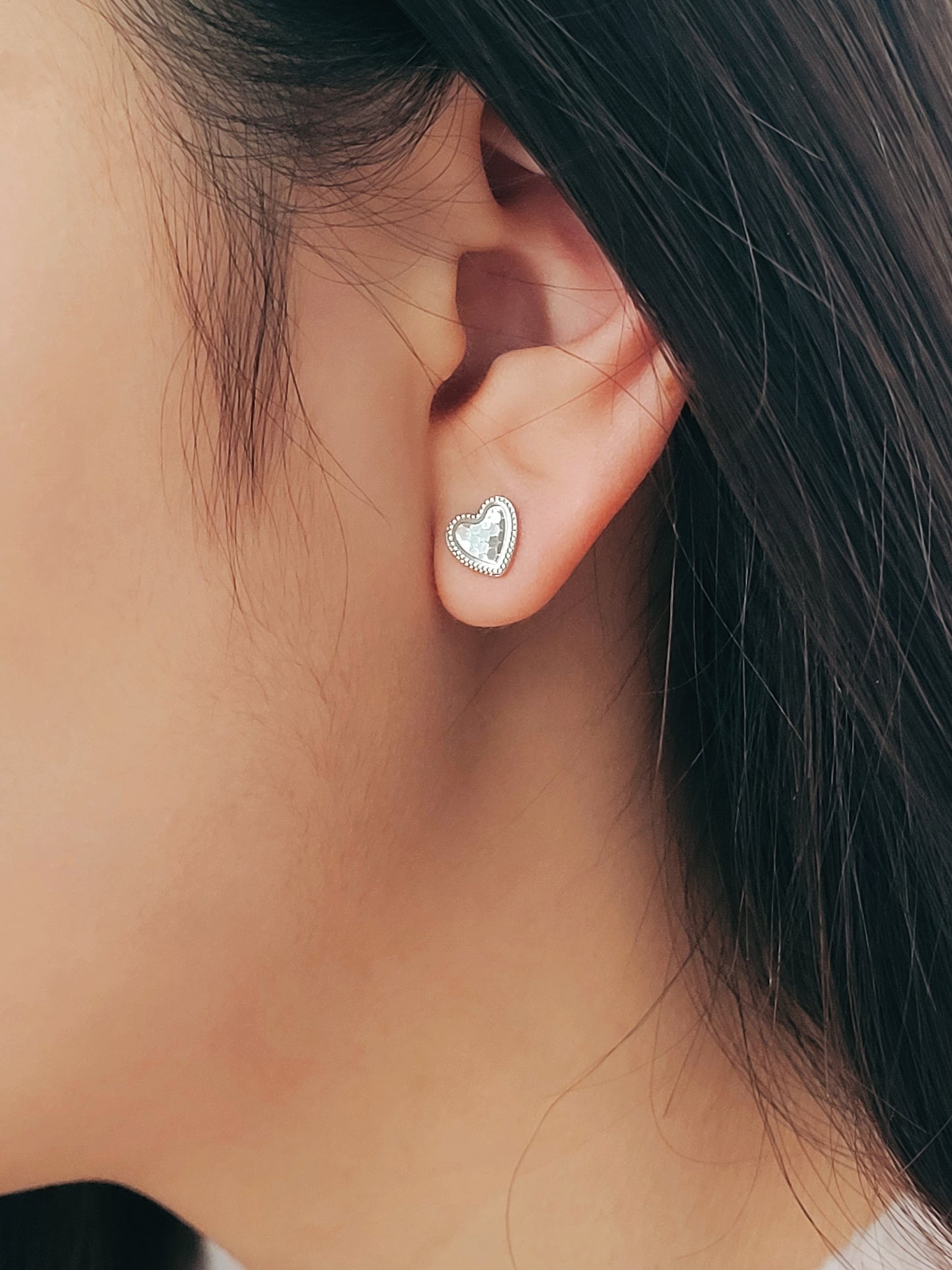 Girl wearing a silver heart stud earrings with subtle textured surface with a fine beaded edge detail.