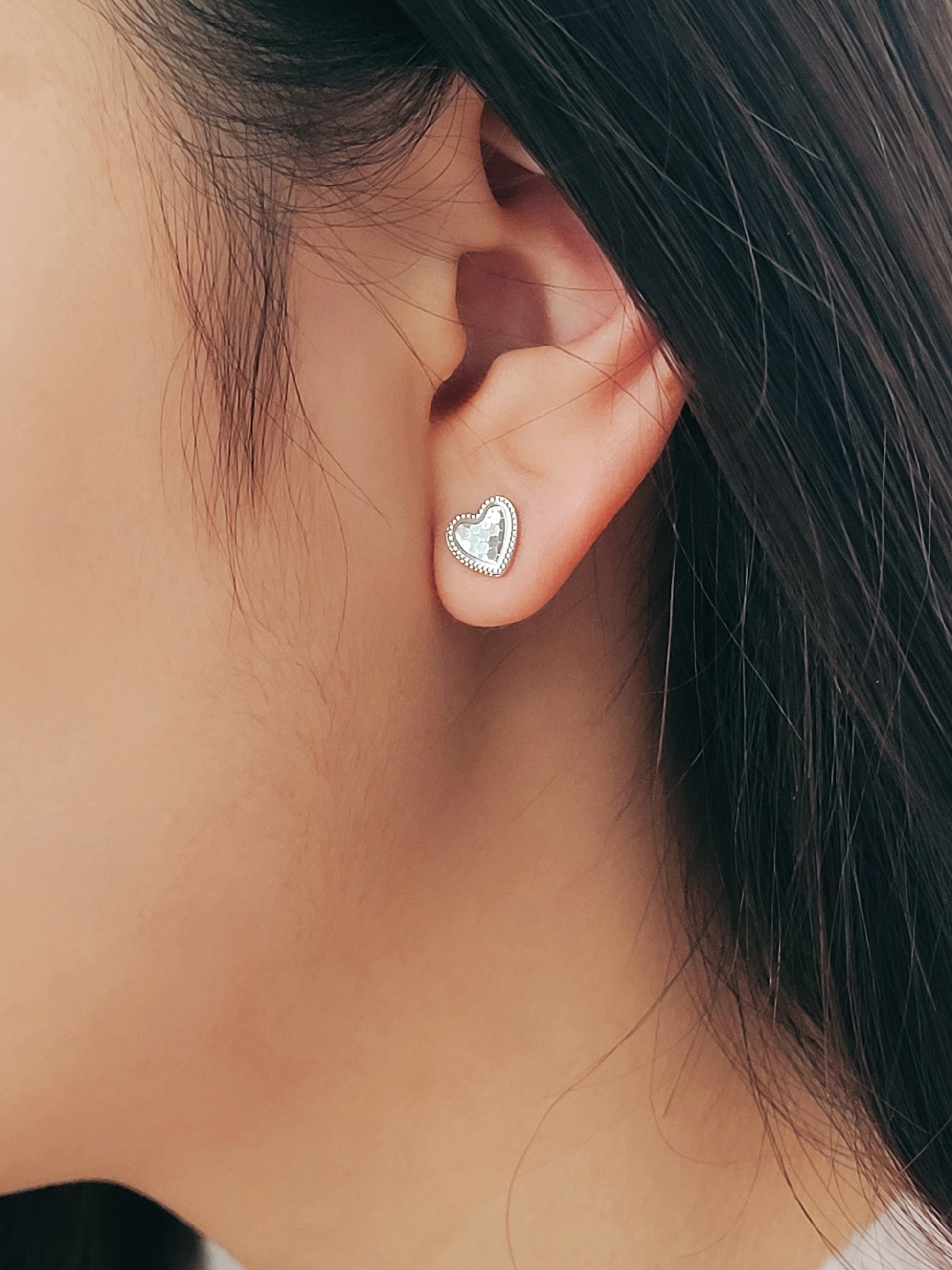 Girl wearing a silver heart stud earrings with subtle textured surface with a fine beaded edge detail.