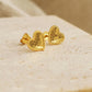 A pair of gold heart earrings feature a textured surface with a fine beaded edge detail displayed on a stone surface.