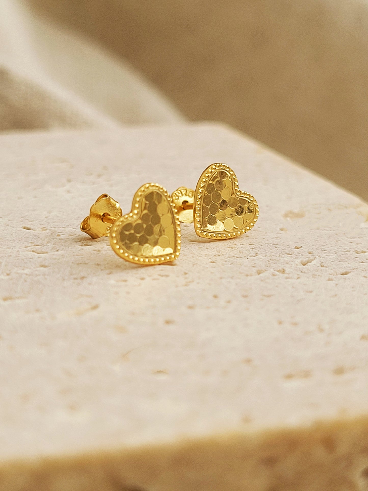 A pair of gold heart earrings feature a textured surface with a fine beaded edge detail displayed on a stone surface.