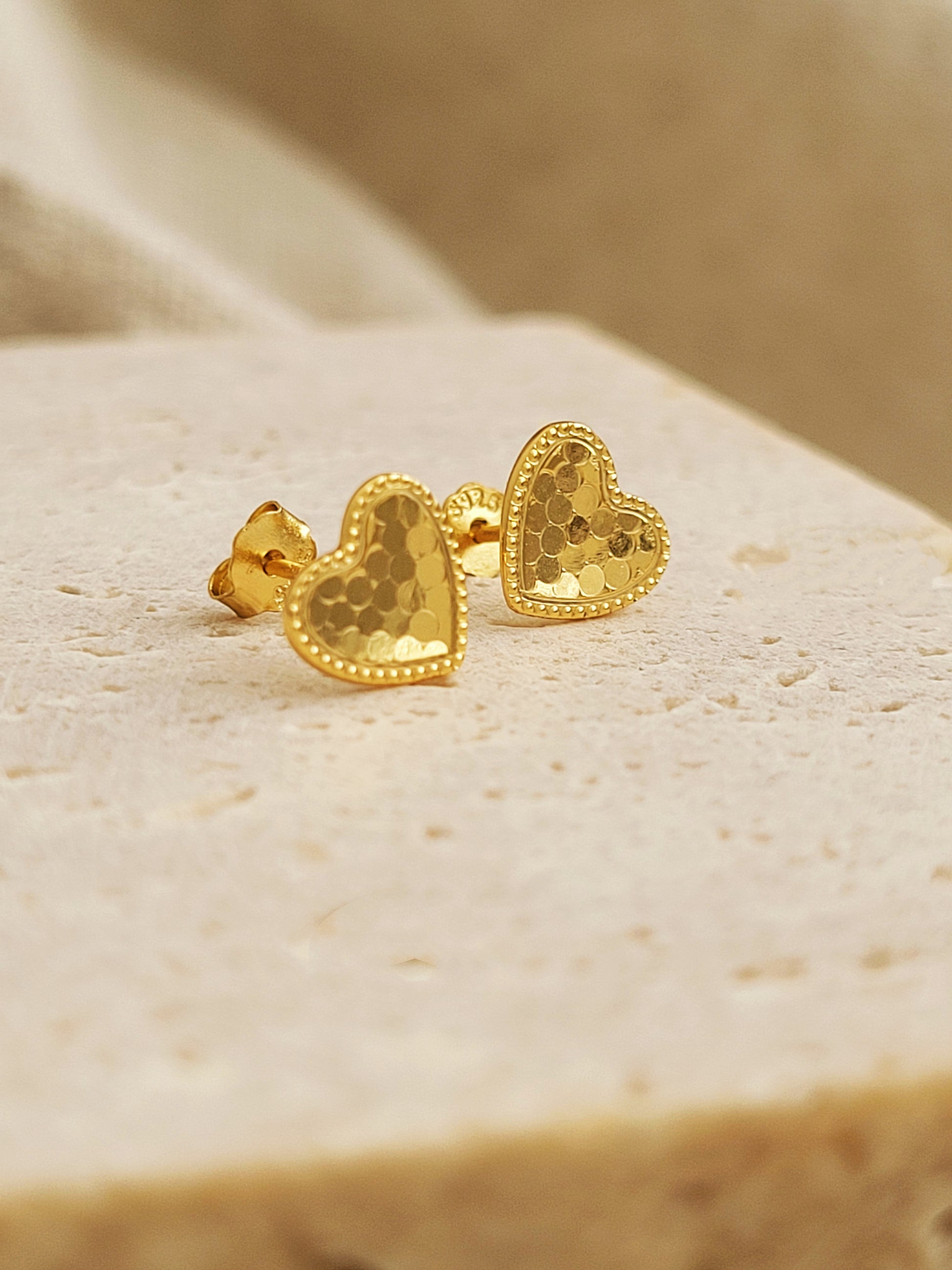 A pair of gold heart earrings feature a textured surface with a fine beaded edge detail displayed on a stone surface.