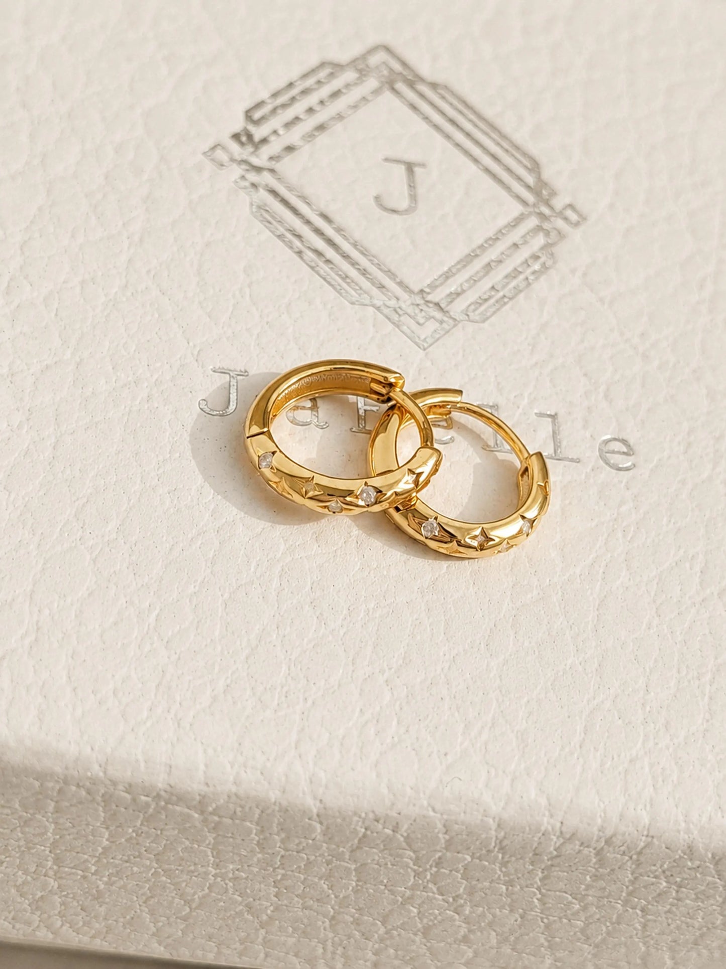 Pair of 18k gold-plated star huggie hoop earrings with cubic zirconia set on a textured beige jewelry box featuring an JaBelle logo.