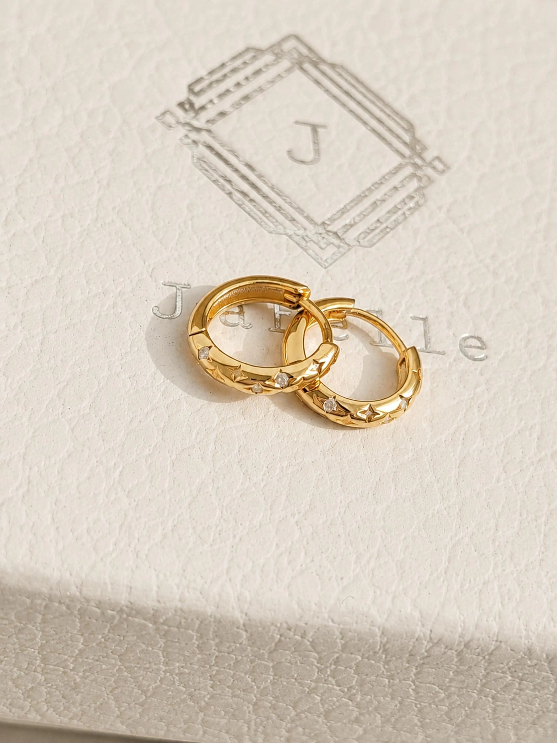 Pair of 18k gold-plated star huggie hoop earrings with cubic zirconia set on a textured beige jewelry box featuring an JaBelle logo.