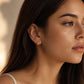 A close-up side profile of a woman with long dark brown hair wearing a tiny gold hoop earring, modern and stylish jewellery, set against a warm beige background with soft shadows.