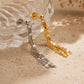A pair of dangling tassel earrings, one in polished gold and one in silver, displayed on a textured beige surface next to a clear glass bowl.