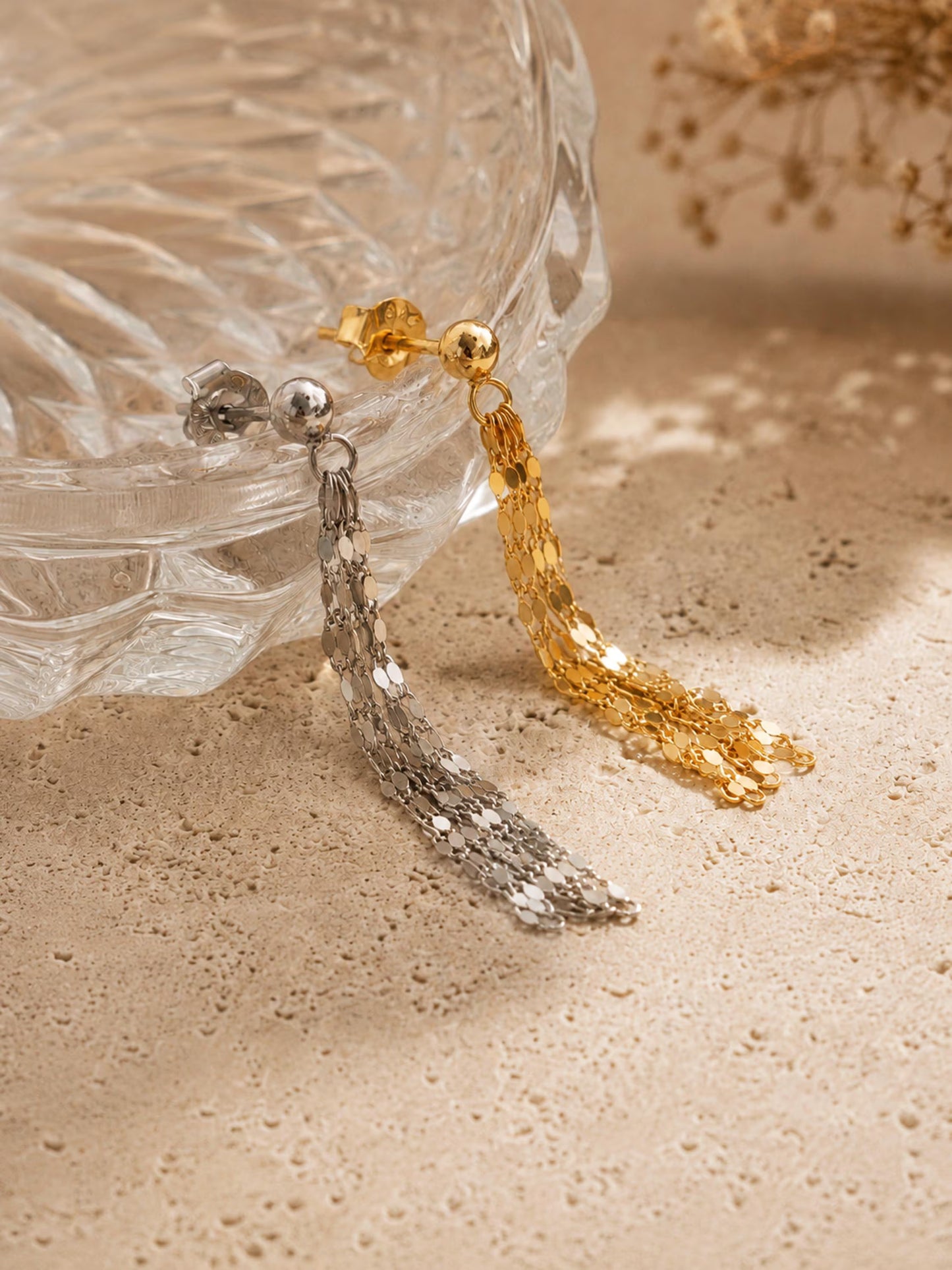 A pair of dangling tassel earrings, one in polished gold and one in silver, displayed on a textured beige surface next to a clear glass bowl.