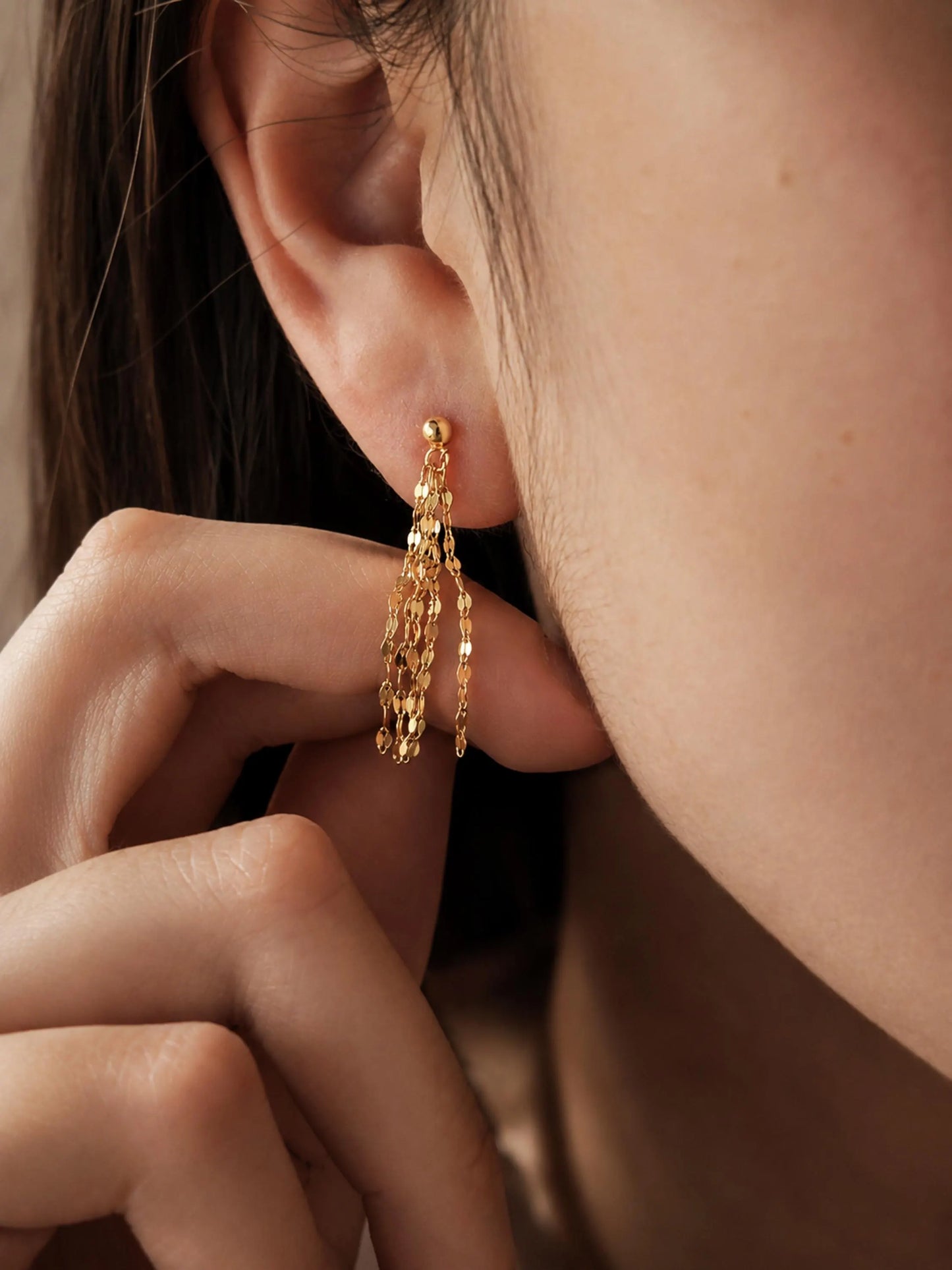 A close-up of a model's ear wearing a delicate gold tassel chain earring with dangling chain and bead fringe crafted in 18k gold plated sterling silver.