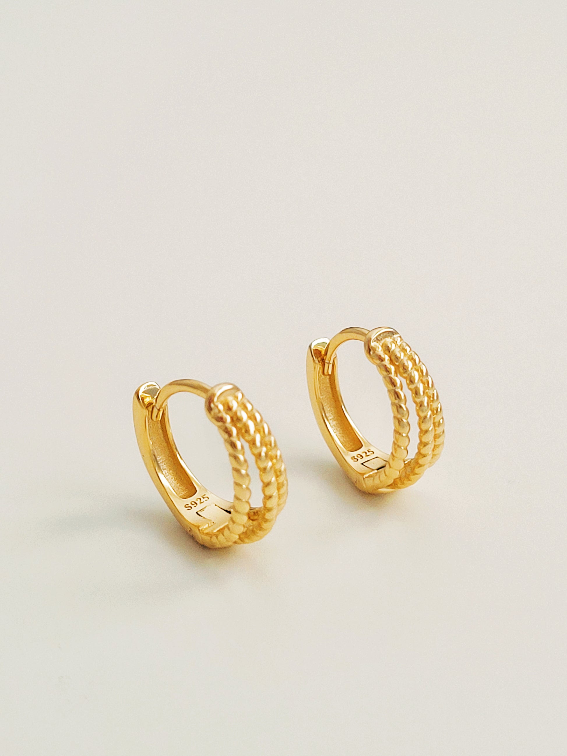 Close up of a pair of minimalist triple hoop twisted huggie earrings crafted in 18k gold plated sterling silver, displayed on a white surface showcasing their details and elegance.