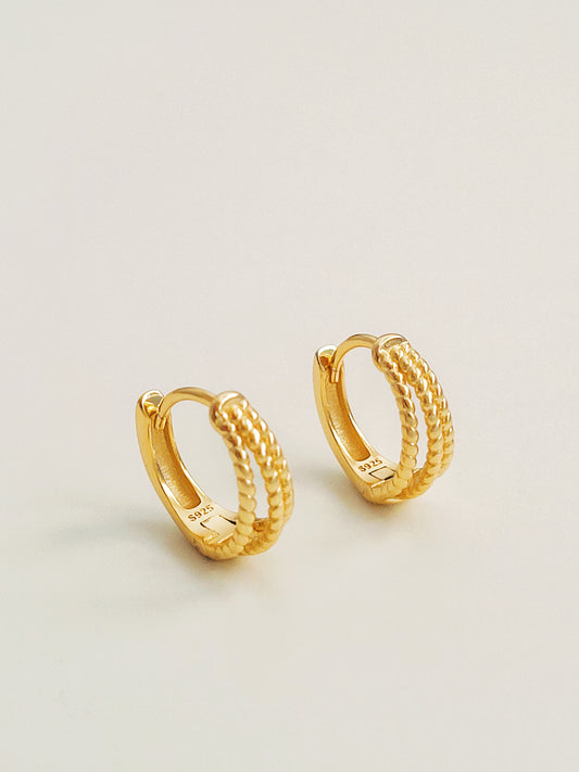 Close up of a pair of minimalist triple hoop twisted huggie earrings crafted in 18k gold plated sterling silver, displayed on a white surface showcasing their details and elegance.