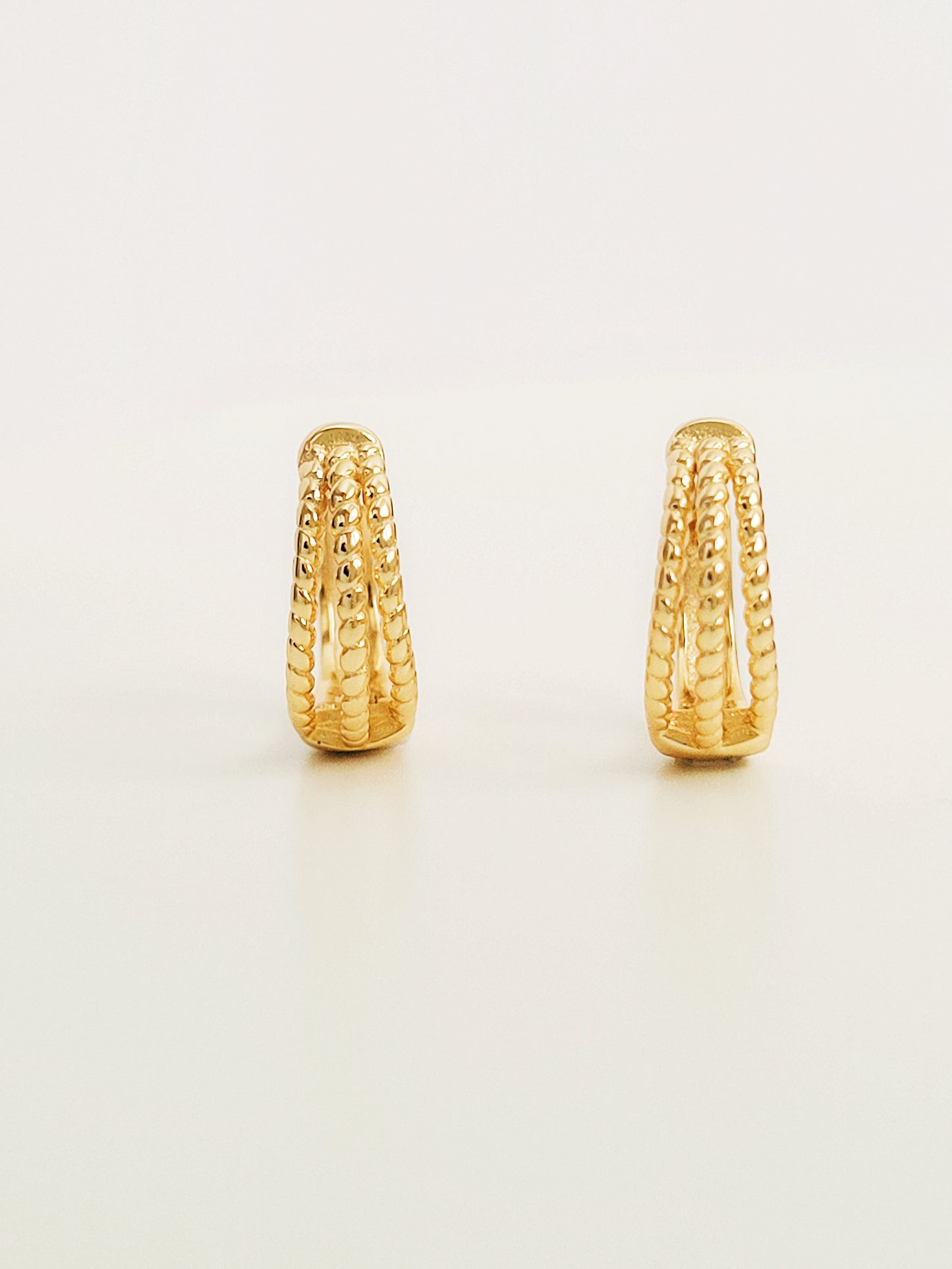 Close up of a pair of minimalist triple hoop twisted huggie earrings crafted in 18k gold plated sterling silver, displayed on a white surface showcasing their details and elegance.