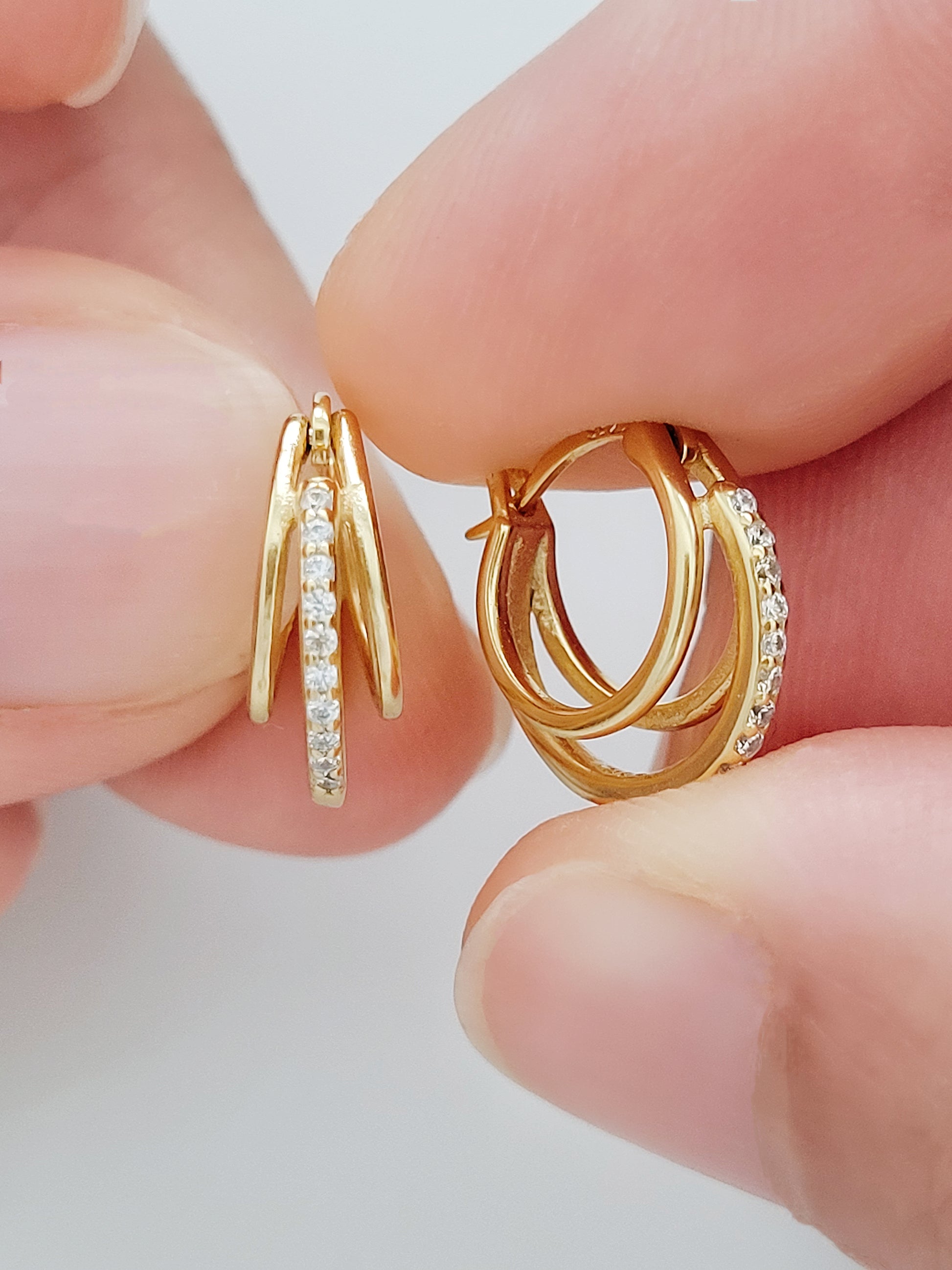 A pair of dazzling triple gold hoop earrings, intricately detailed with sparkling stones, held by a woman's hand, exuding bold luxury and eye-catching brilliance.