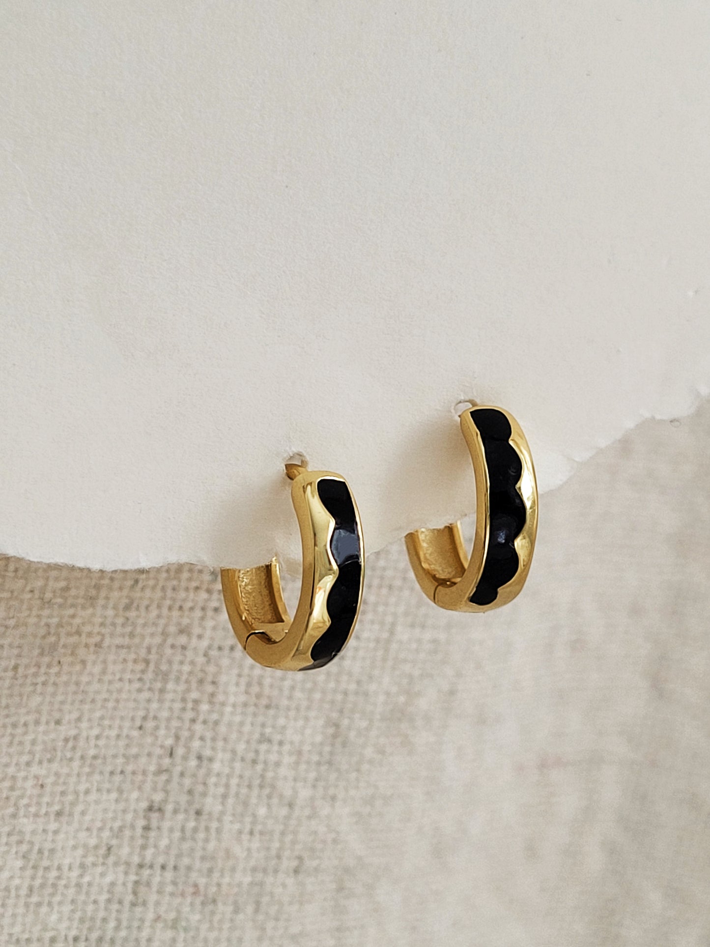 Gold hoop earrings with wavy black enamel on a textured beige surface