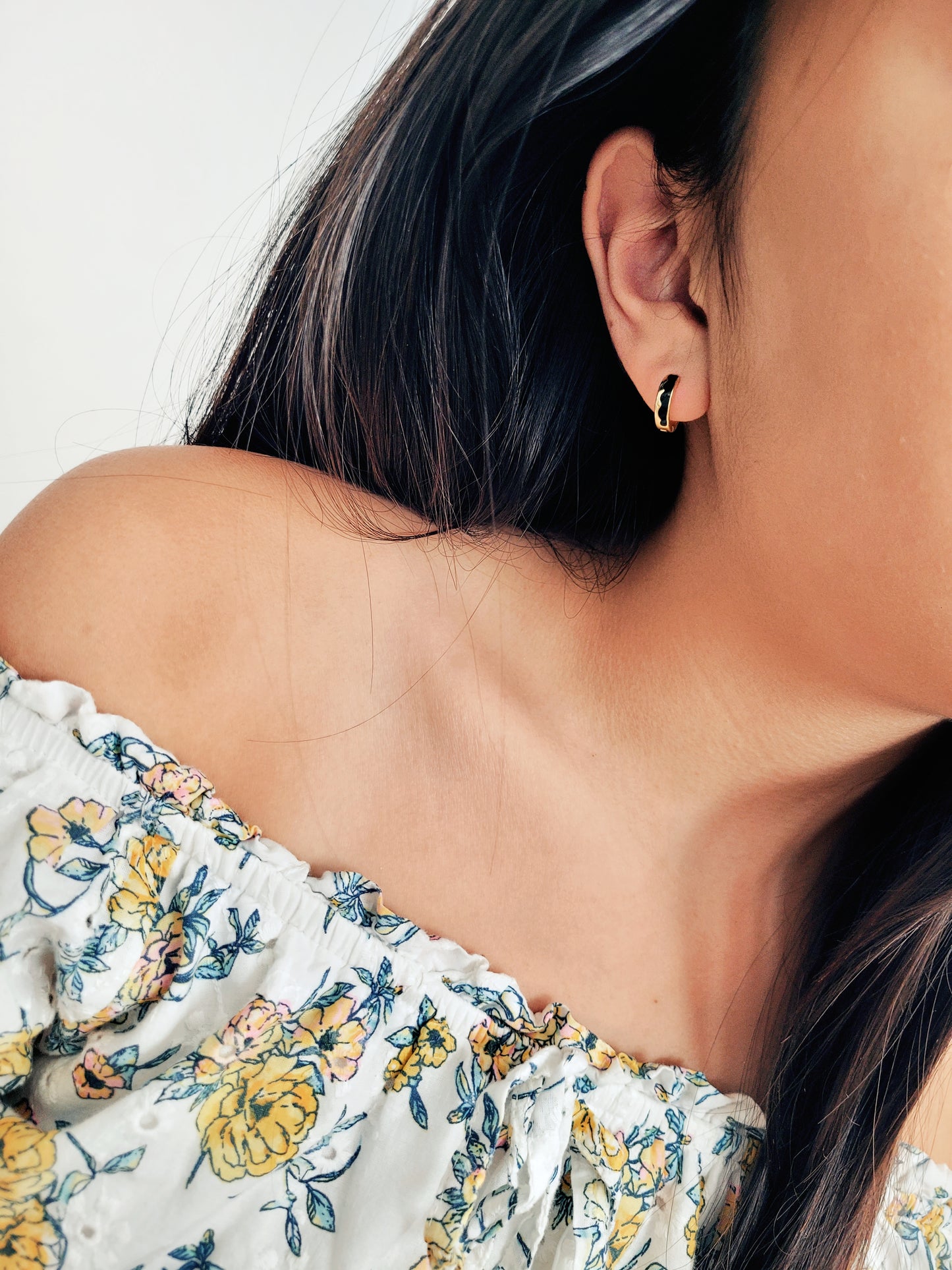 Close-up of a person wearing a floral off-shoulder top with an edgy black enamel huggie earring.