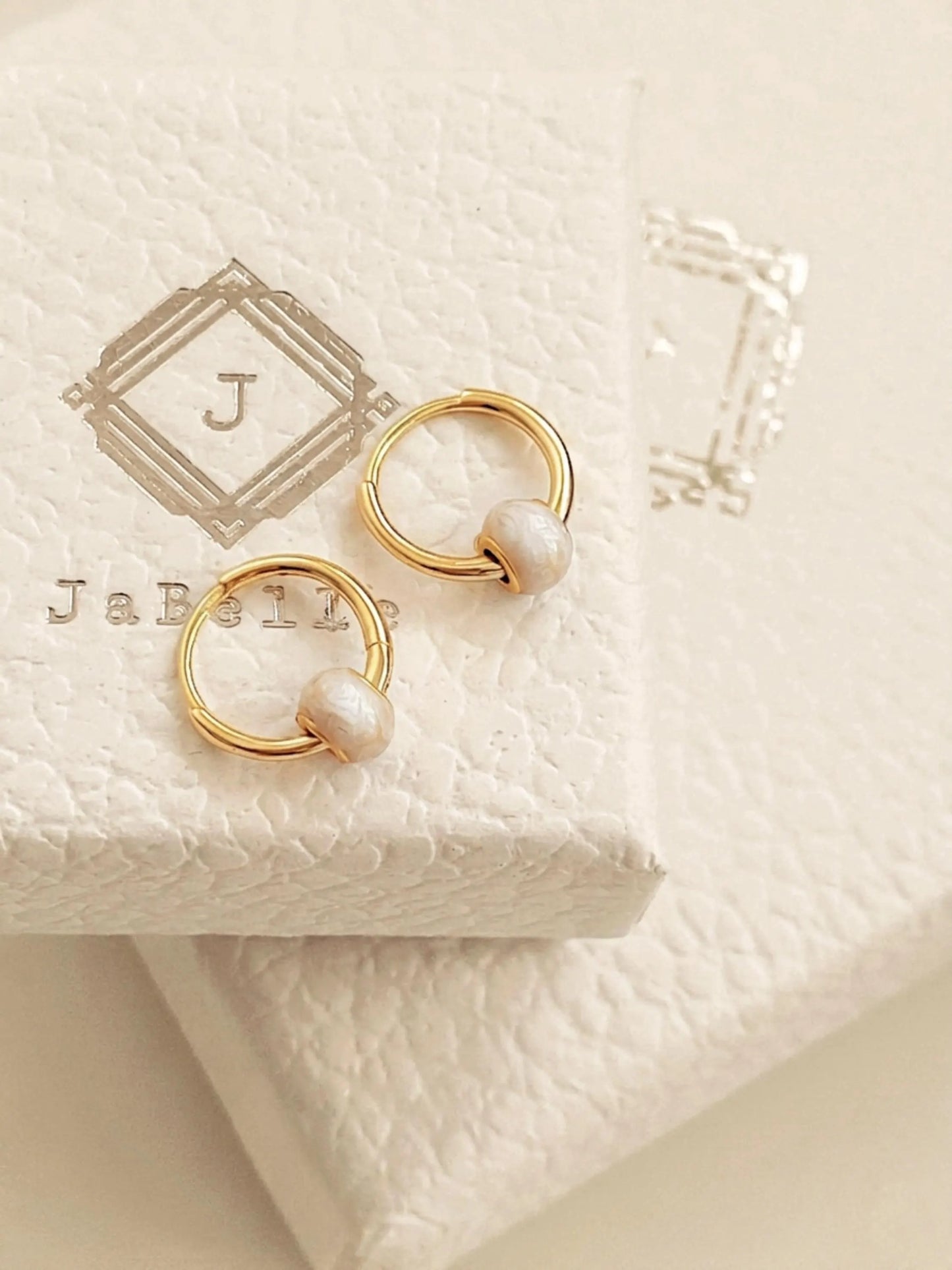 A pair of dainty small gold hoop earrings with white enamel bead displayed on JaBelle Jewellery box.