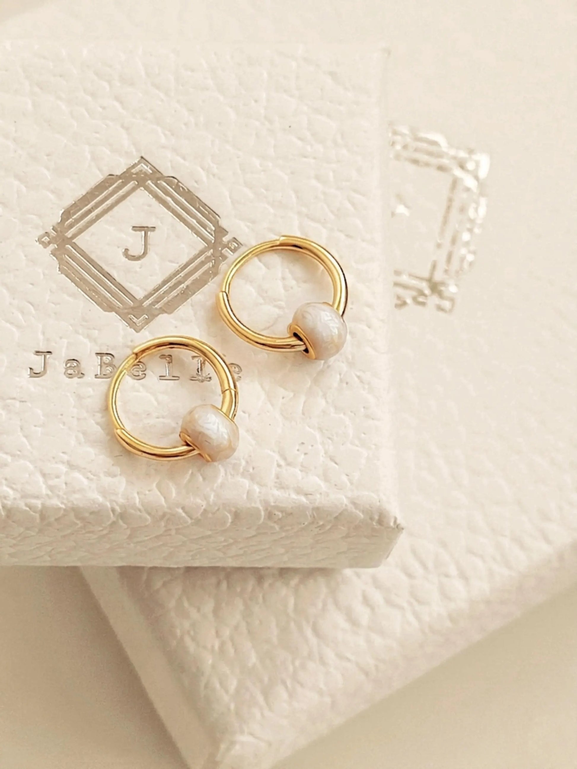 A pair of dainty small gold hoop earrings with white enamel bead displayed on JaBelle Jewellery box.