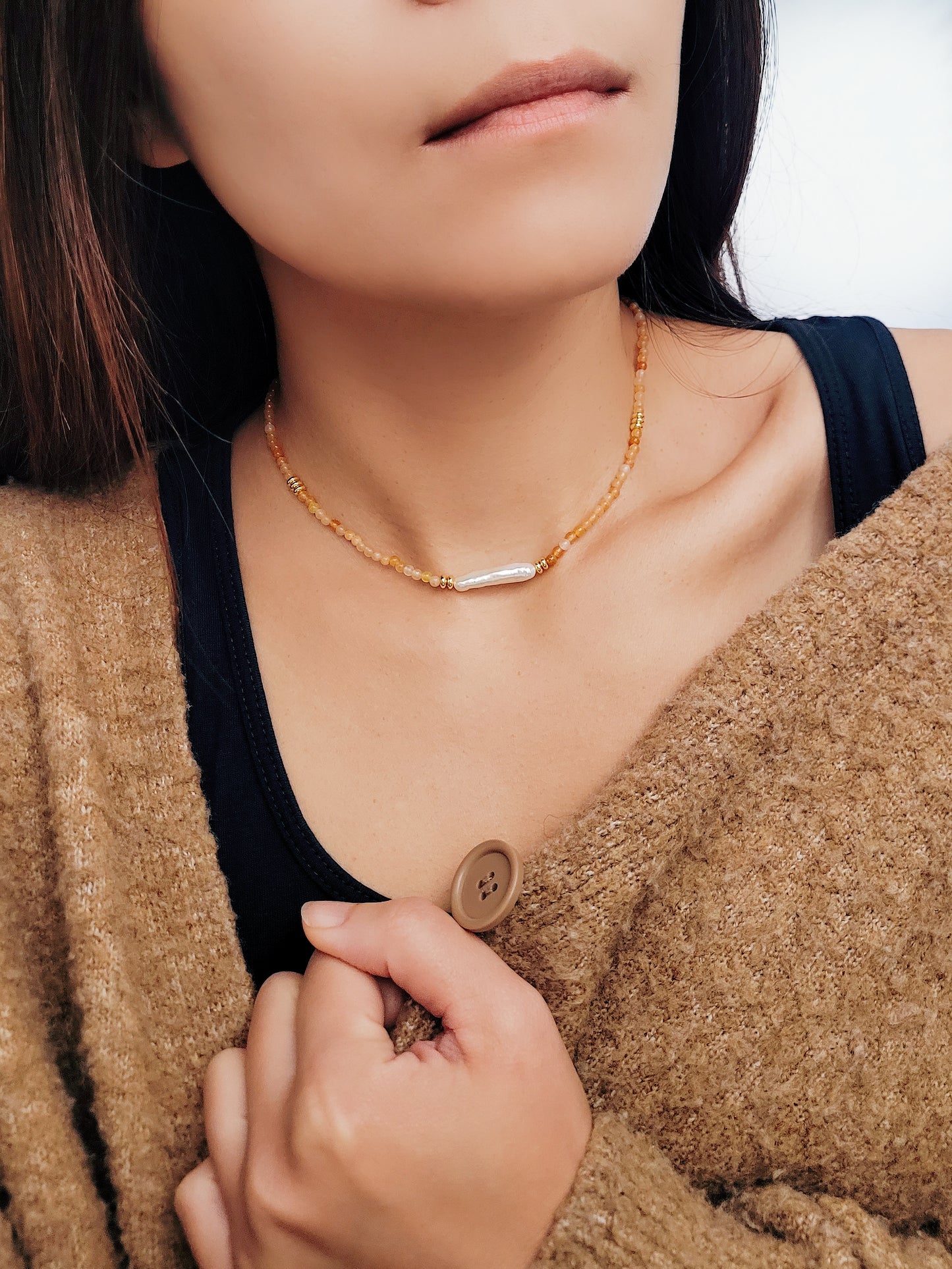 Close-up of a woman wearing an orange agate beaded necklace with a central baroque pearl. The necklace features small, round orange beads accented by gold elements and a single elongated, irregular-shaped white pearl in the center. 