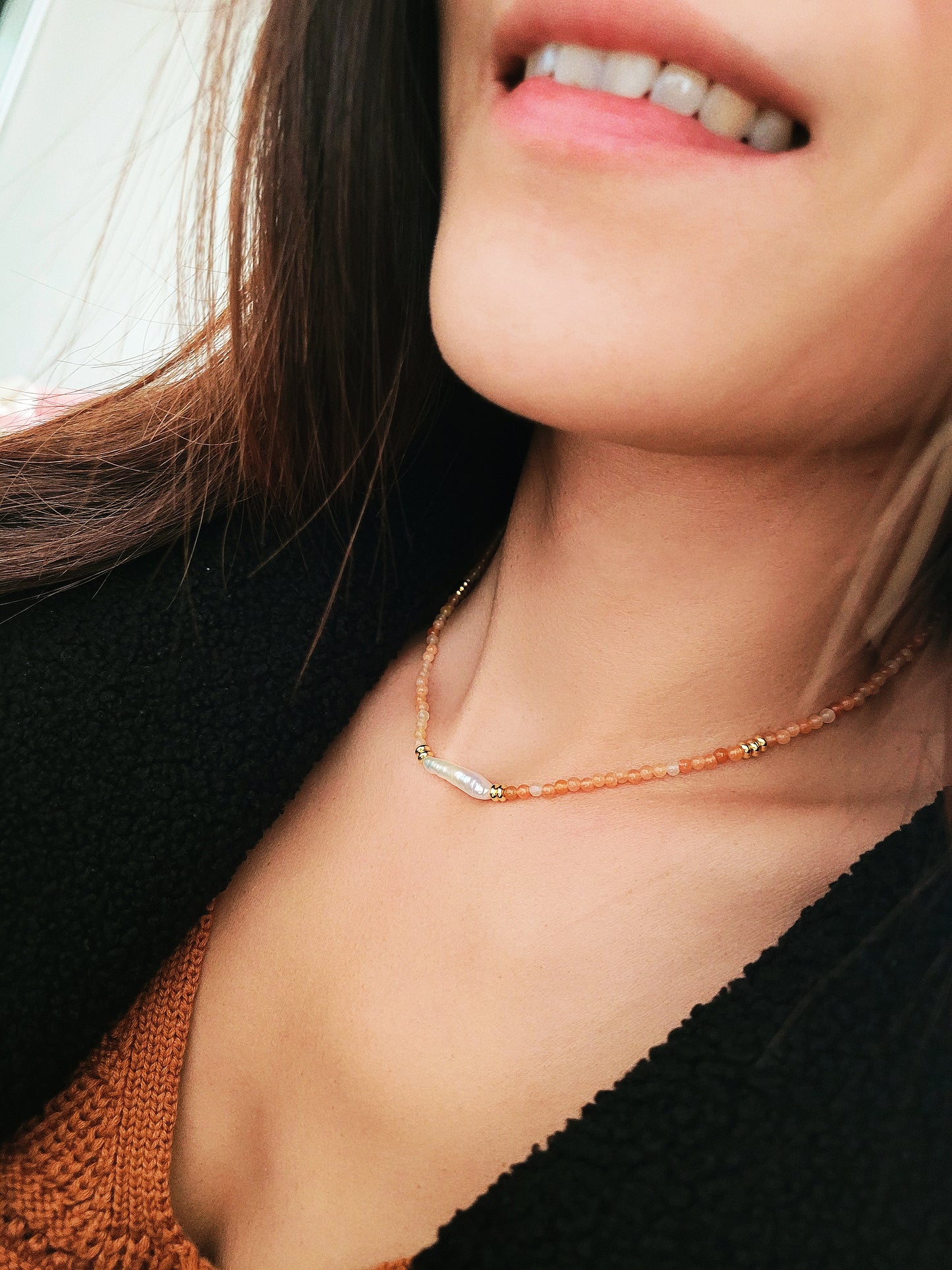 Close-up of a woman wearing an orange agate beaded necklace with a central baroque pearl. The necklace features small, round orange beads accented by gold elements and a single elongated, irregular-shaped white pearl in the center. 