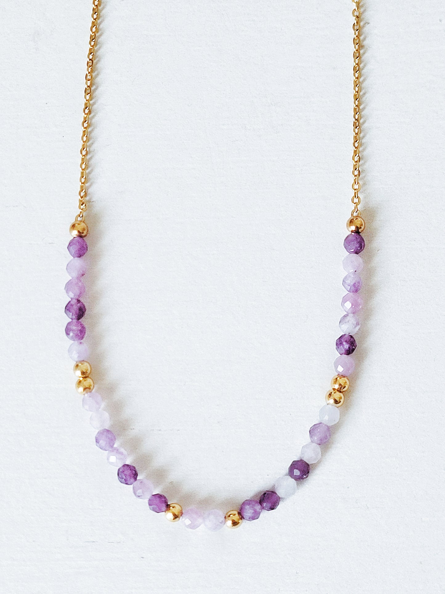 A delicate amethyst beaded necklace featuring small faceted amethyst stones in varying shades of purple, interspersed with gold accents. The beads are arranged in a gradient, transitioning from light to deep purple.