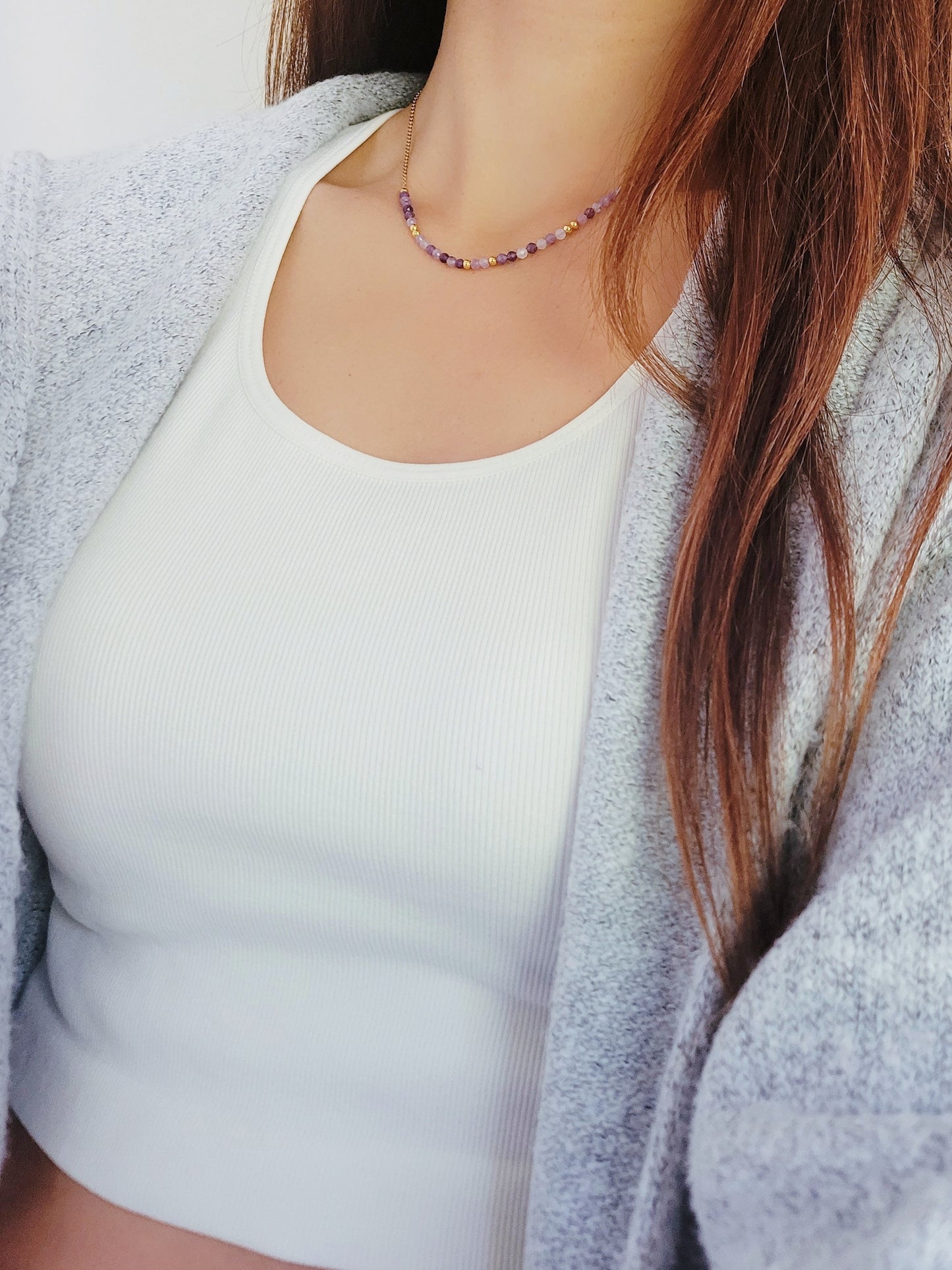 A close-up of a woman wearing a dainty amethyst beaded necklace with faceted purple stones, accented by small gold beads. The necklace with a simple gold chain extending from both sides having purple hues creating a subtle yet stylish look.