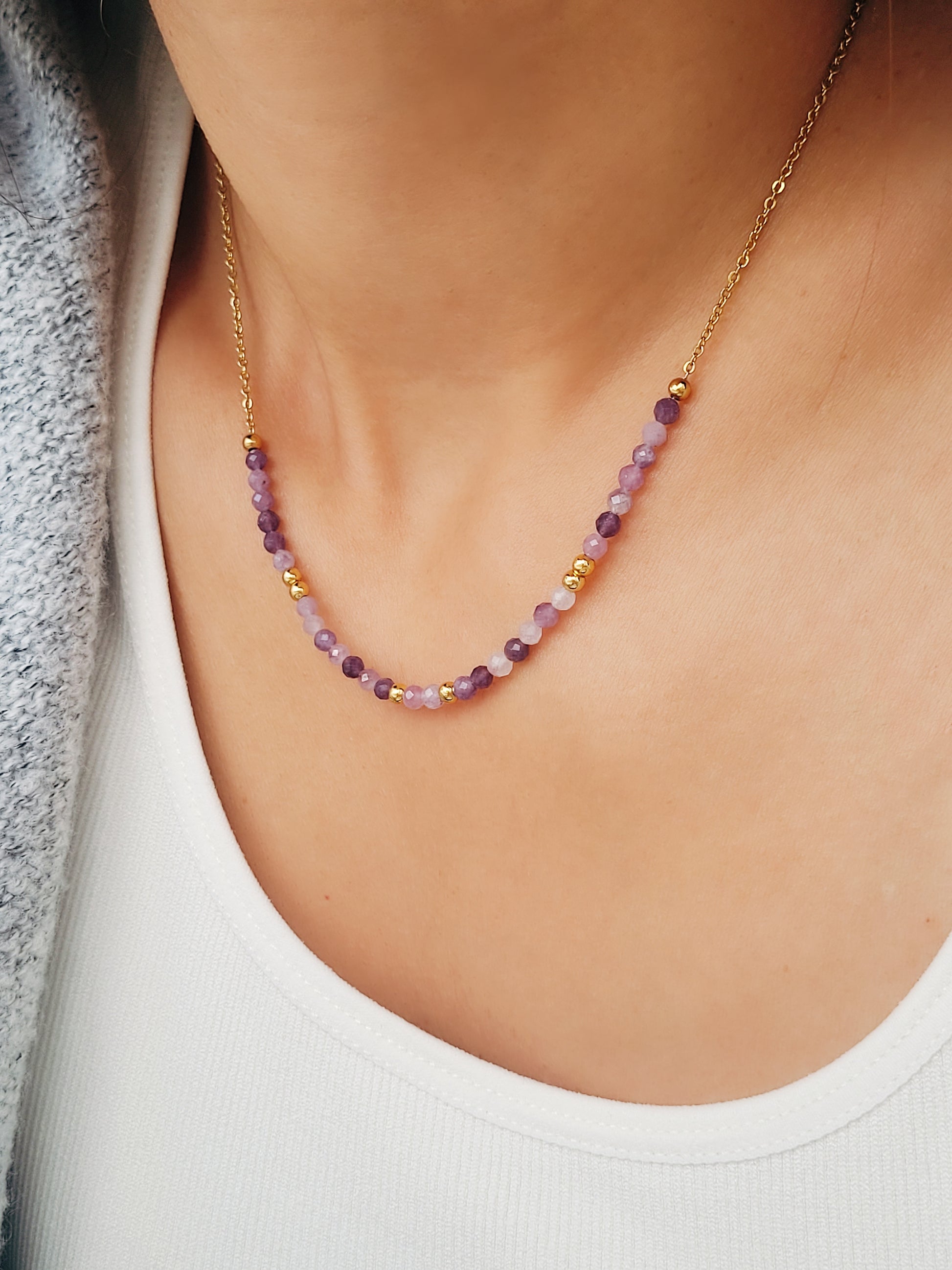 A close-up of a woman wearing a dainty amethyst beaded necklace with faceted purple stones, accented by small gold beads. The necklace with a simple gold chain extending from both sides having purple hues creating a subtle yet stylish look.