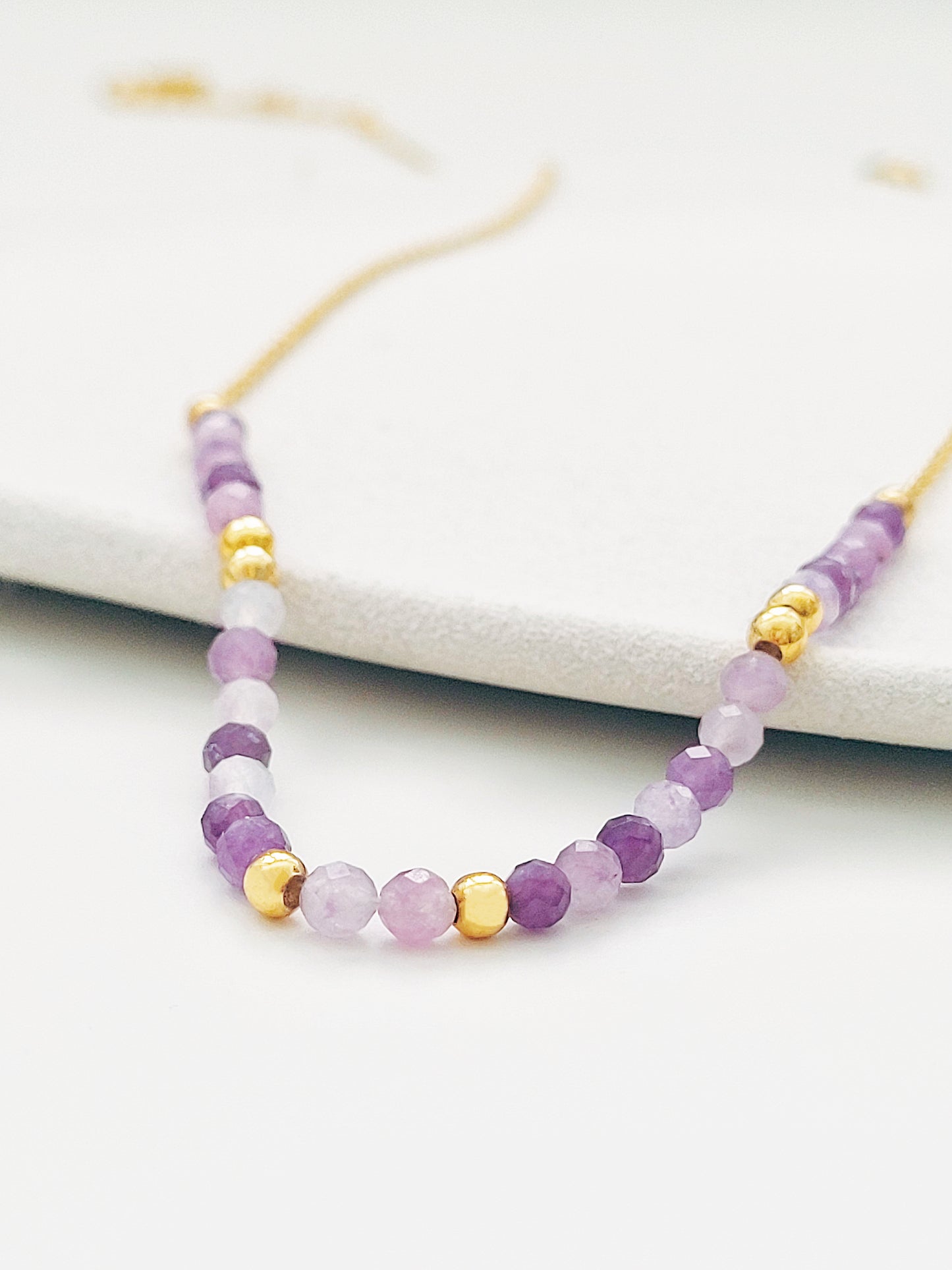 A delicate amethyst beaded necklace featuring small faceted amethyst stones in varying shades of purple, interspersed with gold accents. The beads are arranged in a gradient, transitioning from light to deep purple.