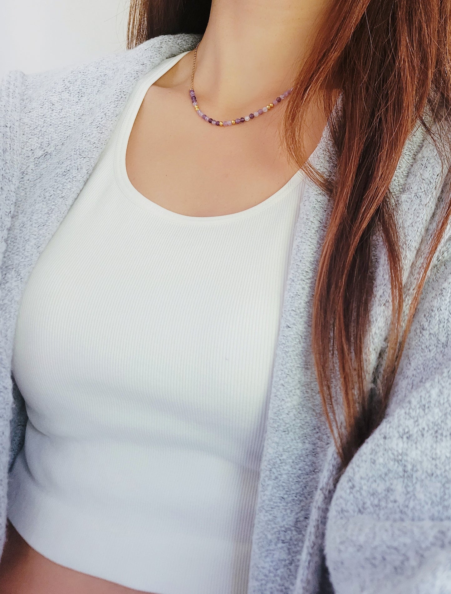 A close-up of a woman wearing a dainty amethyst beaded necklace with faceted purple stones, accented by small gold beads. The necklace with a simple gold chain extending from both sides having purple hues creating a subtle yet stylish look.