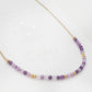 A delicate amethyst beaded necklace featuring small faceted amethyst stones in varying shades of purple, interspersed with gold accents. The beads are arranged in a gradient, transitioning from light to deep purple.
