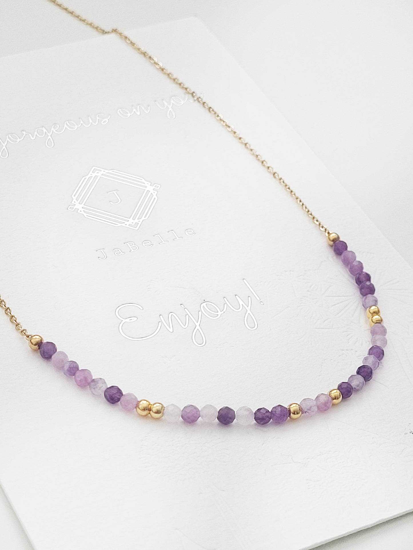 A delicate amethyst beaded necklace featuring small faceted amethyst stones in varying shades of purple, interspersed with gold accents. The beads are arranged in a gradient, transitioning from light to deep purple.