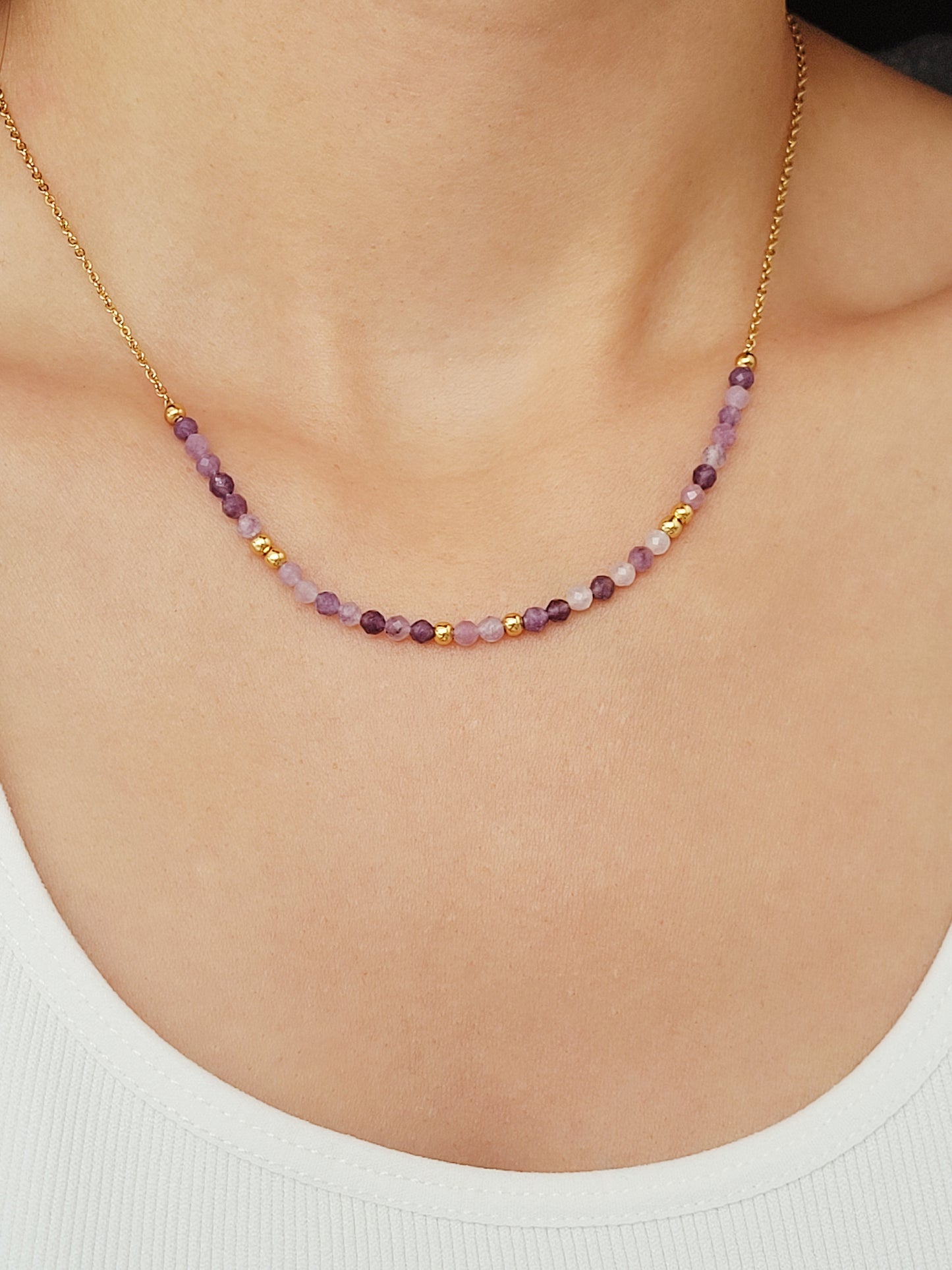 A close-up of a woman wearing a dainty amethyst beaded necklace with faceted purple stones, accented by small gold beads. The necklace with a simple gold chain extending from both sides having purple hues creating a subtle yet stylish look.