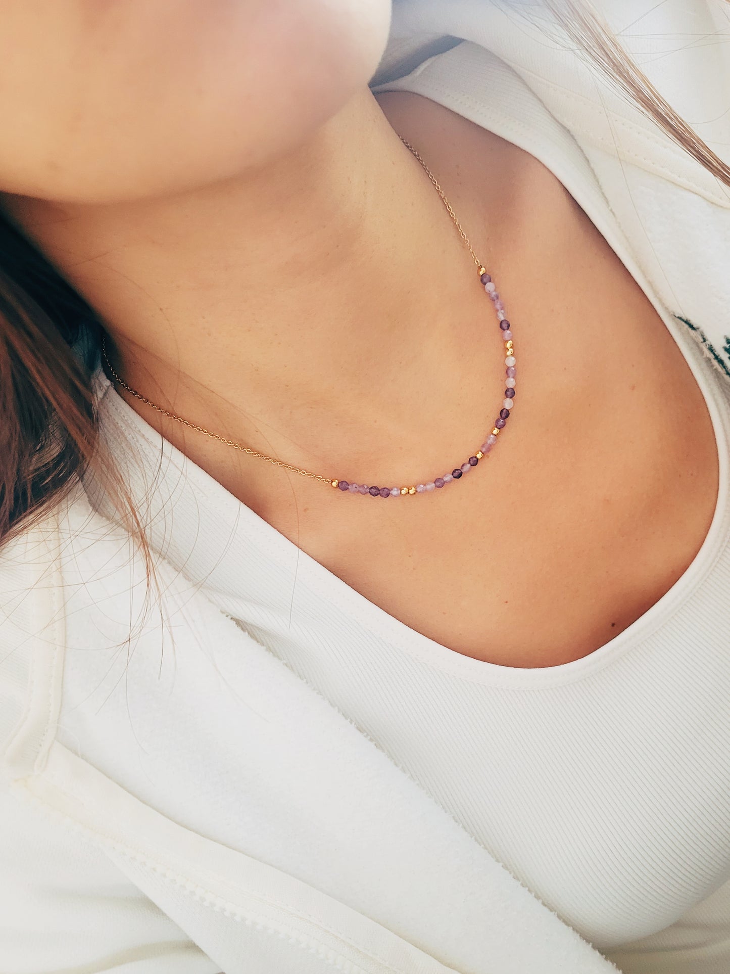 A woman wearing a dainty amethyst beaded necklace with faceted purple stones, accented by small gold beads. The necklace with a simple gold chain extending from both sides having purple hues creating a subtle yet stylish look.