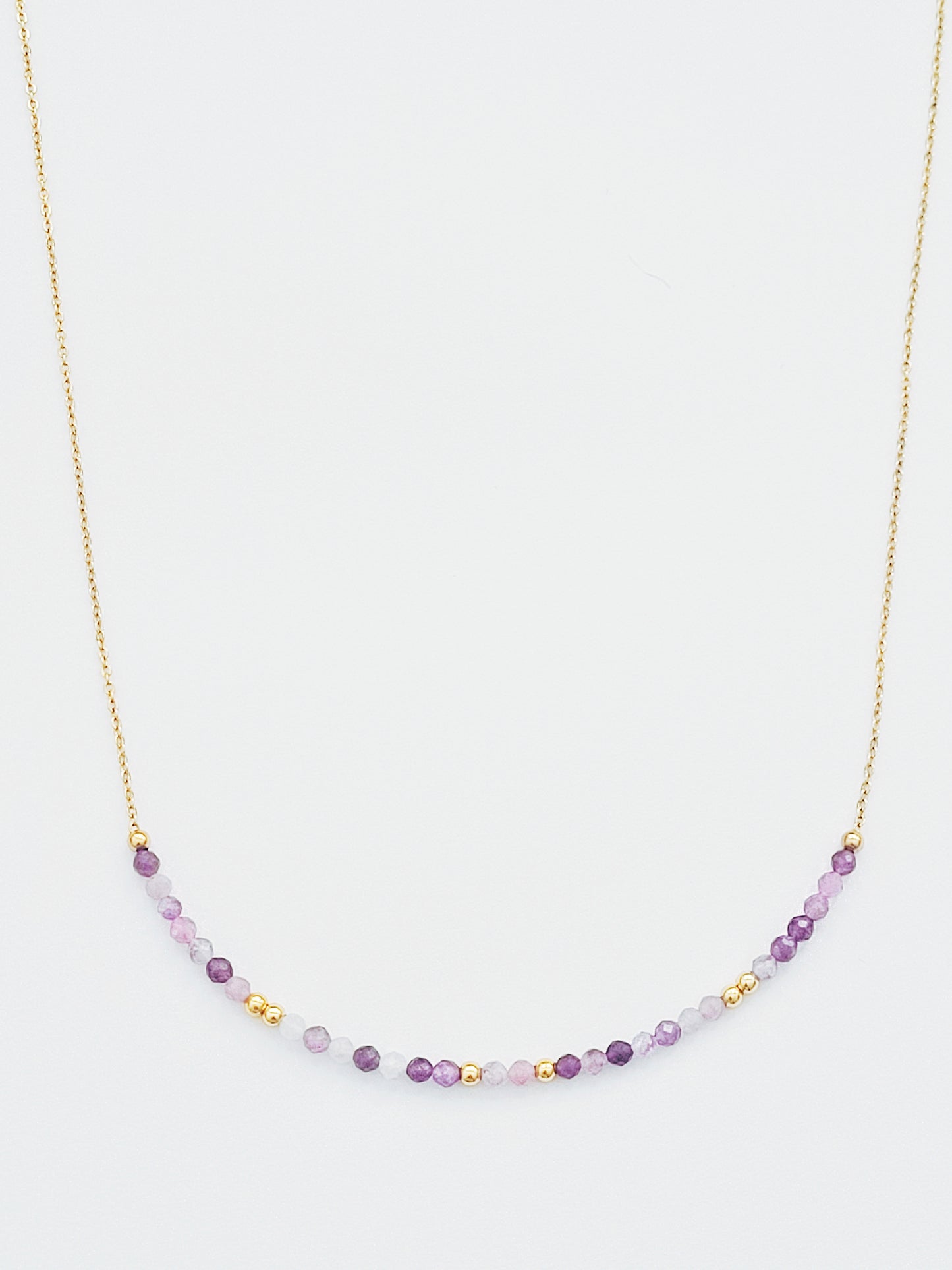 A delicate amethyst beaded necklace featuring small faceted amethyst stones in varying shades of purple, interspersed with gold accents. The beads are arranged in a gradient, transitioning from light to deep purple.