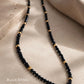 A close-up shot of a delicate black gemstone necklace laid out gracefully on a textured beige stone tile. The necklace features faceted, sparkling black spinel beads interspersed with smooth, round 22k gold vermeil accent beads. Warm, soft natural light casts gentle shadows, highlighting the luxury texture. Text overlay on the bottom left reads 'Black Spinel 22K Gold Vermeil'.