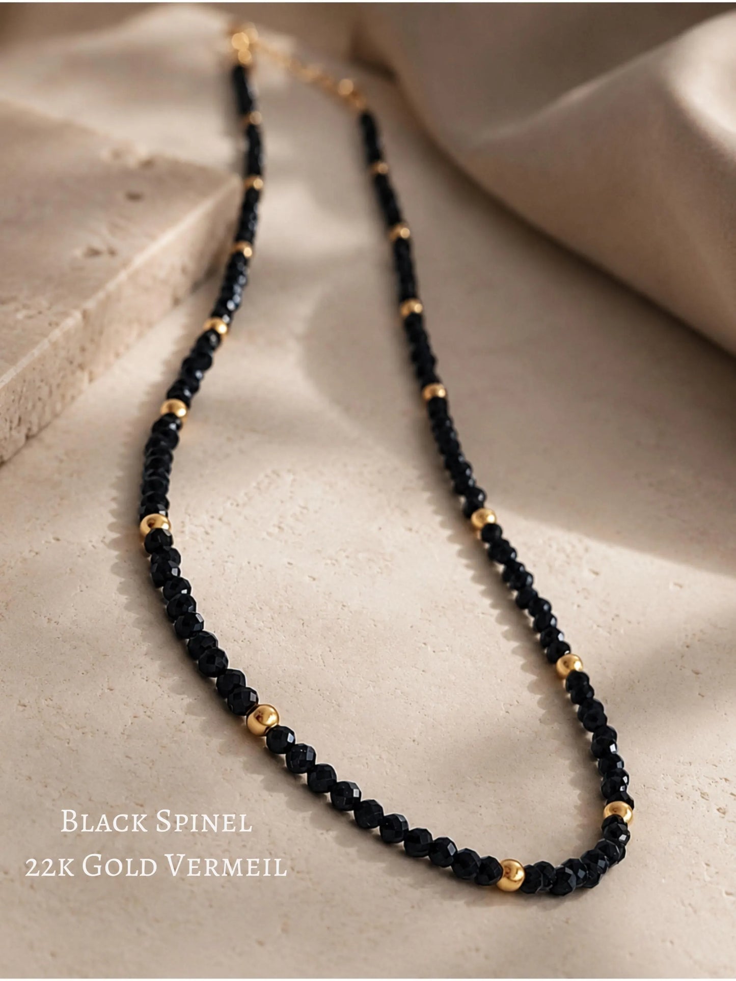 A close-up shot of a delicate black gemstone necklace laid out gracefully on a textured beige stone tile. The necklace features faceted, sparkling black spinel beads interspersed with smooth, round 22k gold vermeil accent beads. Warm, soft natural light casts gentle shadows, highlighting the luxury texture. Text overlay on the bottom left reads 'Black Spinel 22K Gold Vermeil'.