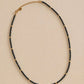 A delicate black spinel choker necklace displayed on a plain surface highlights the shimmering, faceted black gemstone beads and the polished, round 22k gold vermeil accents. The scene is shot in warm, golden lighting in the background.