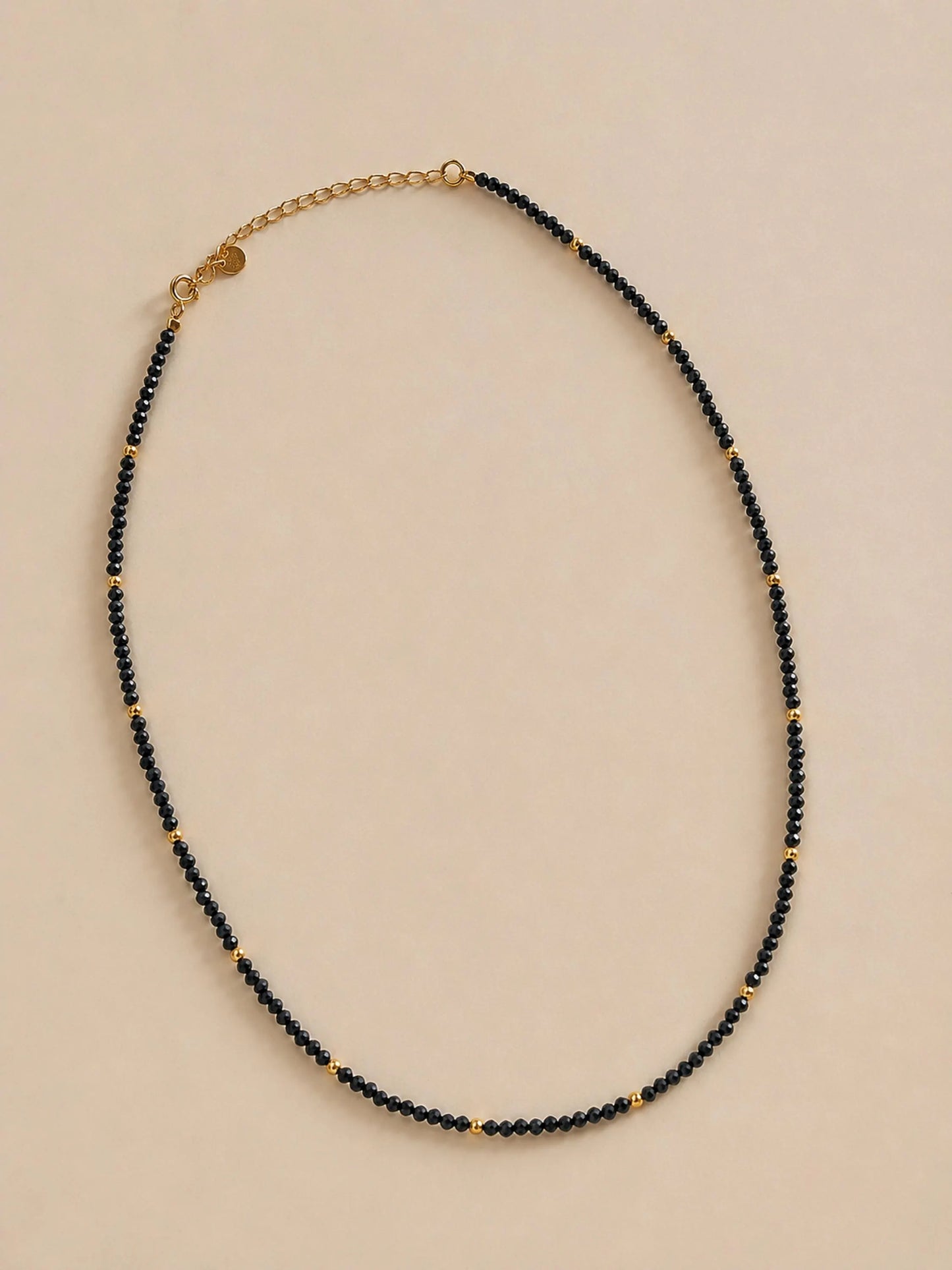 A delicate black spinel choker necklace displayed on a plain surface highlights the shimmering, faceted black gemstone beads and the polished, round 22k gold vermeil accents. The scene is shot in warm, golden lighting in the background.
