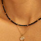 Close-up detail of a layered necklace look on a model's neck. A dainty black spinel choker with small, polished 22k gold vermeil accent beads is paired beautifully with a longer, fine gold chain necklace featuring a round, textured disc pendant. The shot highlights how to style and layer the minimalist beaded jewellery.