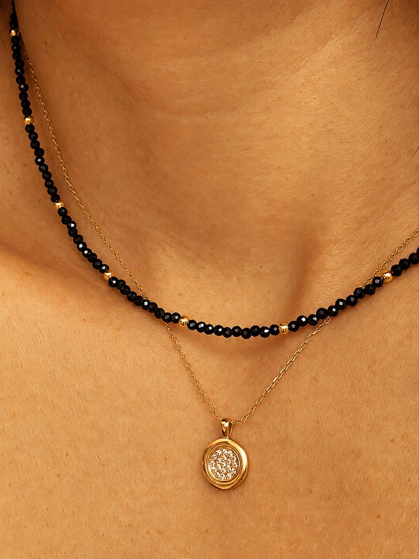 Close-up detail of a layered necklace look on a model's neck. A dainty black spinel choker with small, polished 22k gold vermeil accent beads is paired beautifully with a longer, fine gold chain necklace featuring a round, textured disc pendant. The shot highlights how to style and layer the minimalist beaded jewellery.