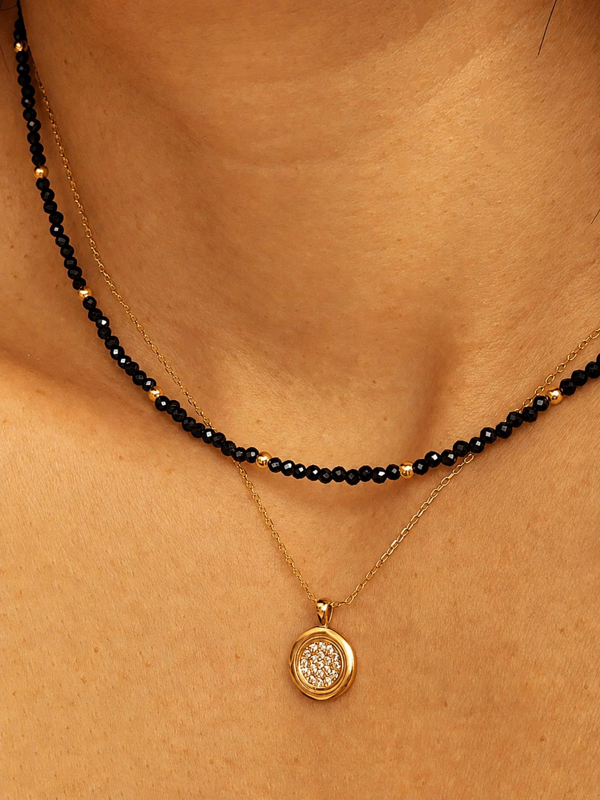 Close-up detail of a layered necklace look on a model's neck. A dainty black spinel choker with small, polished 22k gold vermeil accent beads is paired beautifully with a longer, fine gold chain necklace featuring a round, textured disc pendant. The shot highlights how to style and layer the minimalist beaded jewellery.