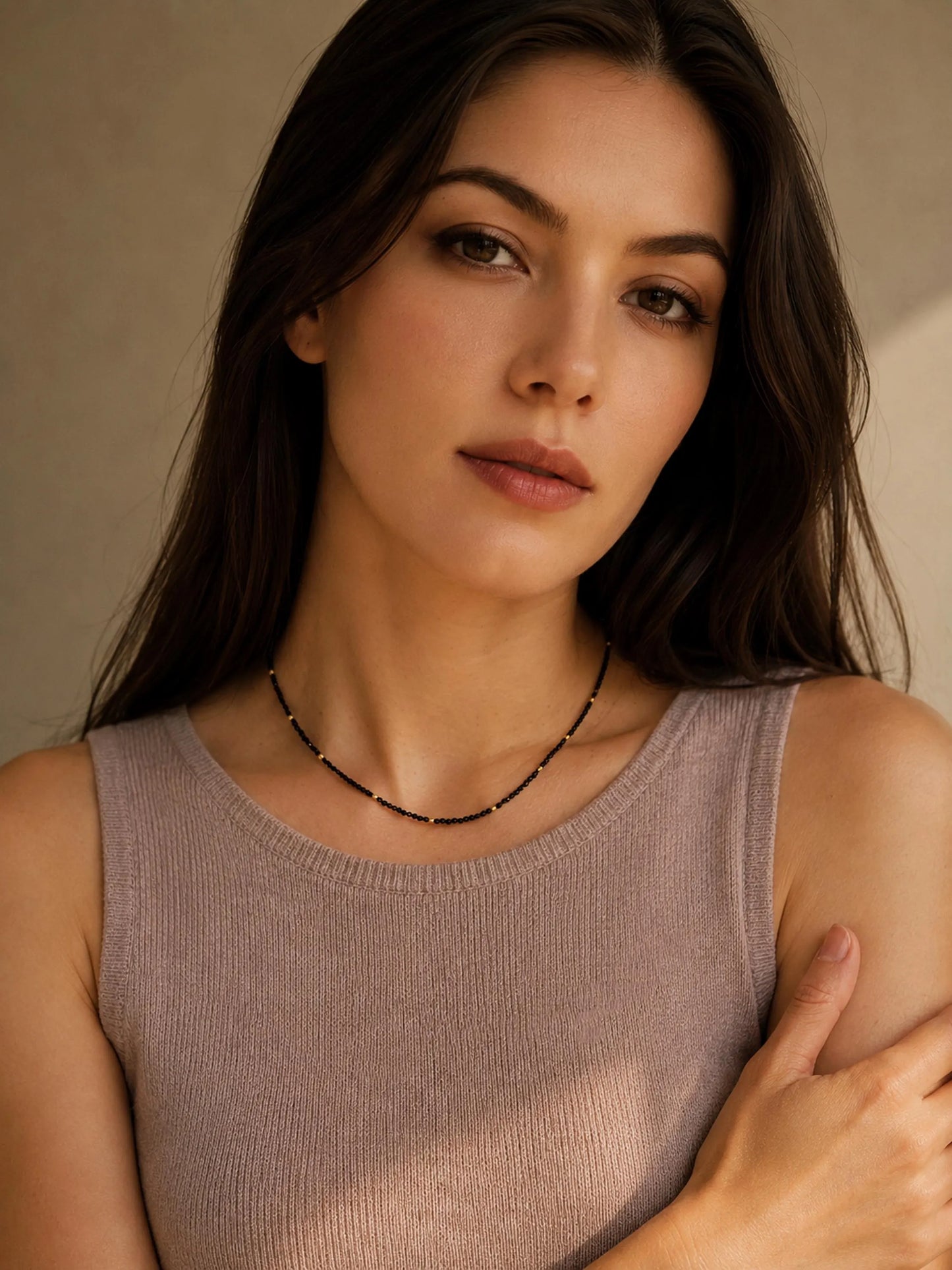 Portrait of a female model with long brunette hair wearing a dainty black spinel choker necklace. The delicate necklace features tiny, faceted black beads accented by small 22k gold vermeil beads, resting gracefully above her collarbone. She is styled in a ribbed, sleeveless mauve-taupe knit top against a warm, neutral background.