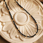 A delicate black spinel and 22k gold vermeil beaded necklace arranged in a neat curve over a round, cream-colored plaster medallion with a raised floral sunburst pattern. Warm, directional sunlight creates crisp shadows across the sculpted background, accentuating the sparkling, faceted texture of the black gemstones.