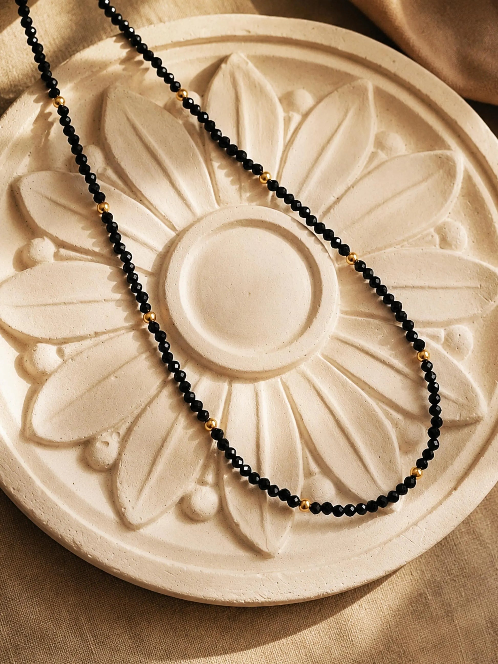 A delicate black spinel and 22k gold vermeil beaded necklace arranged in a neat curve over a round, cream-colored plaster medallion with a raised floral sunburst pattern. Warm, directional sunlight creates crisp shadows across the sculpted background, accentuating the sparkling, faceted texture of the black gemstones.