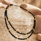 A delicate black spinel choker necklace draped elegantly across a vintage-style, clear cut-glass trinket jar. The double-layered positioning highlights the shimmering, faceted black gemstone beads and the polished, round 22k gold vermeil accents. The scene is shot in warm, golden lighting with soft, draped neutral fabrics in the background.