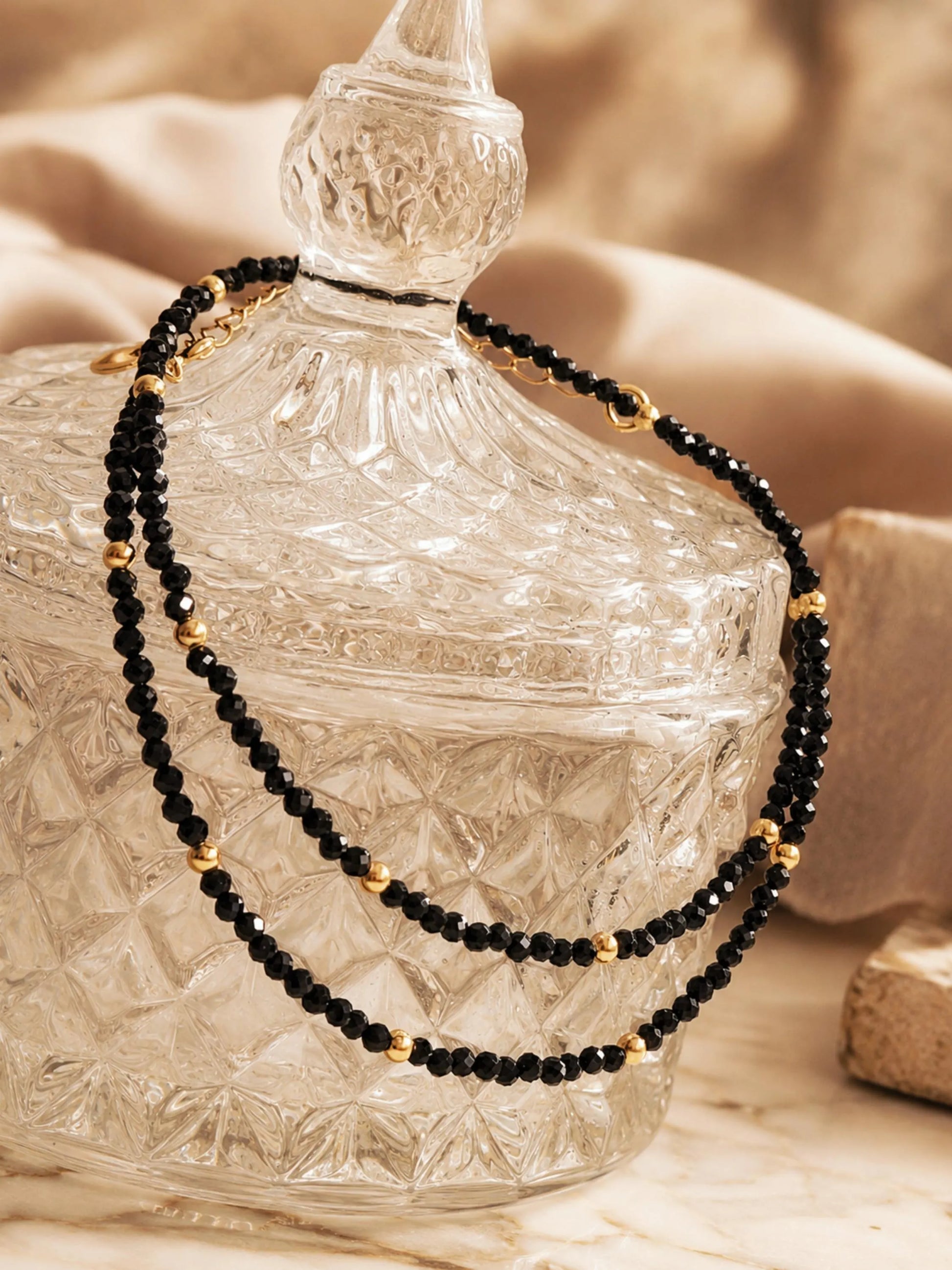 A delicate black spinel choker necklace draped elegantly across a vintage-style, clear cut-glass trinket jar. The double-layered positioning highlights the shimmering, faceted black gemstone beads and the polished, round 22k gold vermeil accents. The scene is shot in warm, golden lighting with soft, draped neutral fabrics in the background.