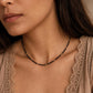 Close-up lifestyle view of a model wearing a dainty black spinel choker necklace with small 22k gold vermeil accent beads. The minimalist beaded necklace rests perfectly on her collarbone, styled alongside a neutral beige top with delicate lace detailing. The shot showcases the elegant scale and fit of the gemstone jewellery on the wrist and neck.