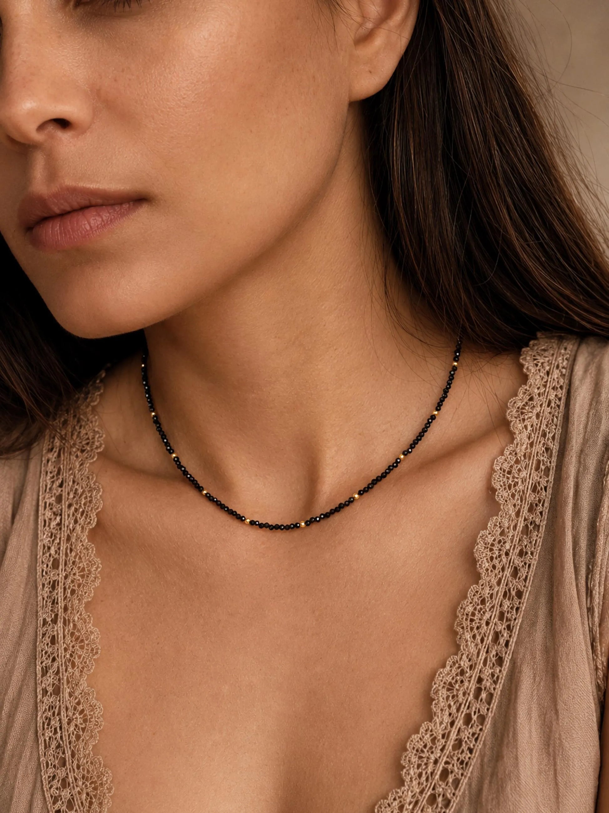 Close-up lifestyle view of a model wearing a dainty black spinel choker necklace with small 22k gold vermeil accent beads. The minimalist beaded necklace rests perfectly on her collarbone, styled alongside a neutral beige top with delicate lace detailing. The shot showcases the elegant scale and fit of the gemstone jewellery on the wrist and neck.